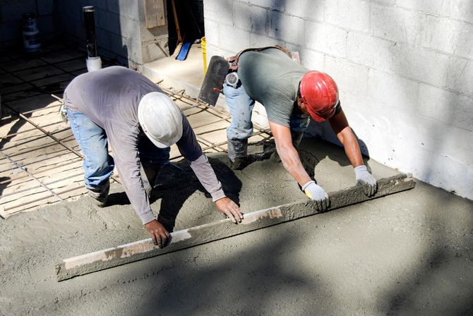Two contractors using a screed board to level a fresh concrete slab.