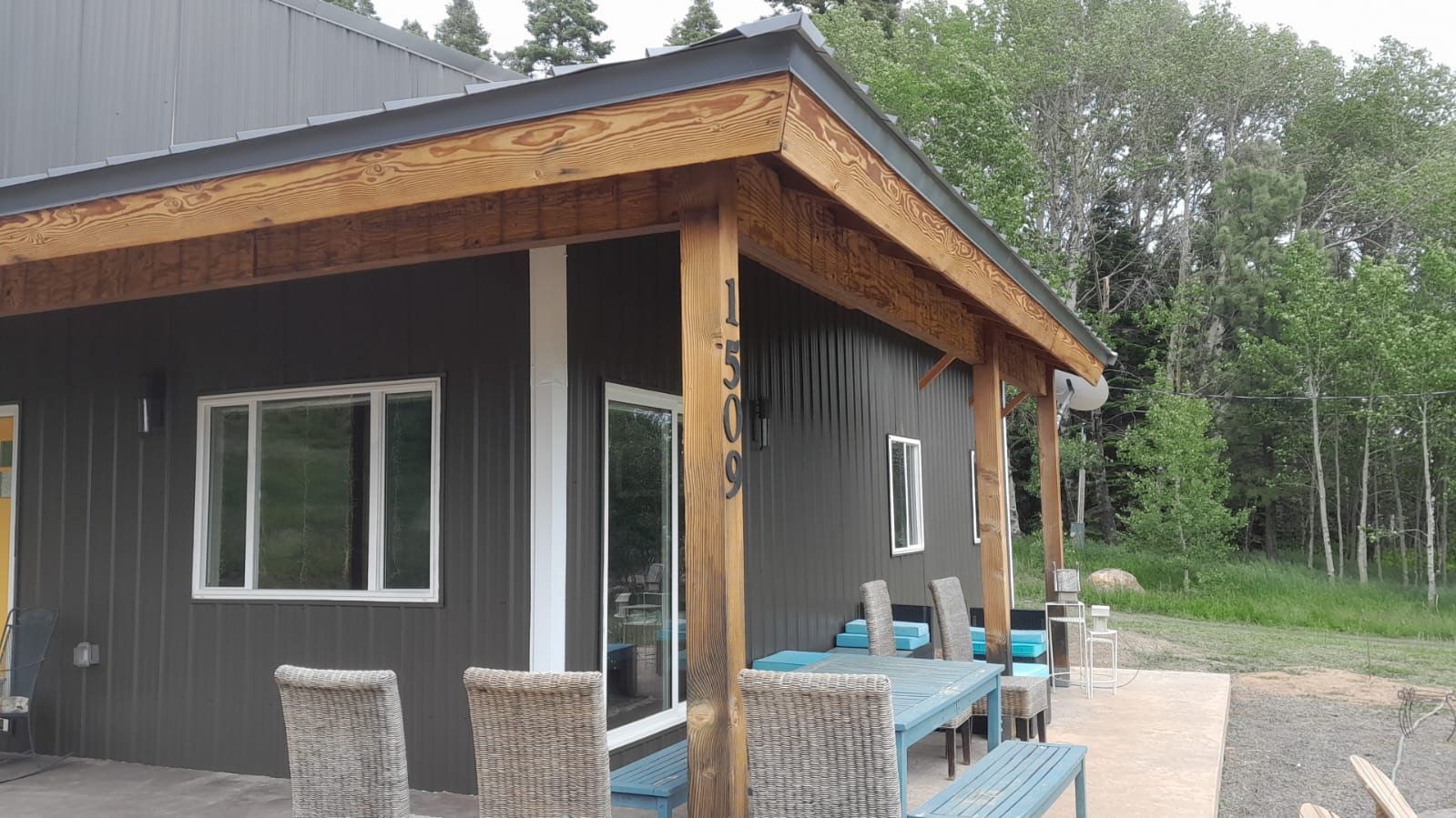 Modern dark-gray house exterior with wood trim, patio chairs, and a blue deck in a wooded setting