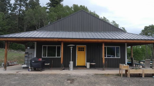 Rustic cabin with black roof, yellow door, and porch furniture surrounded by trees