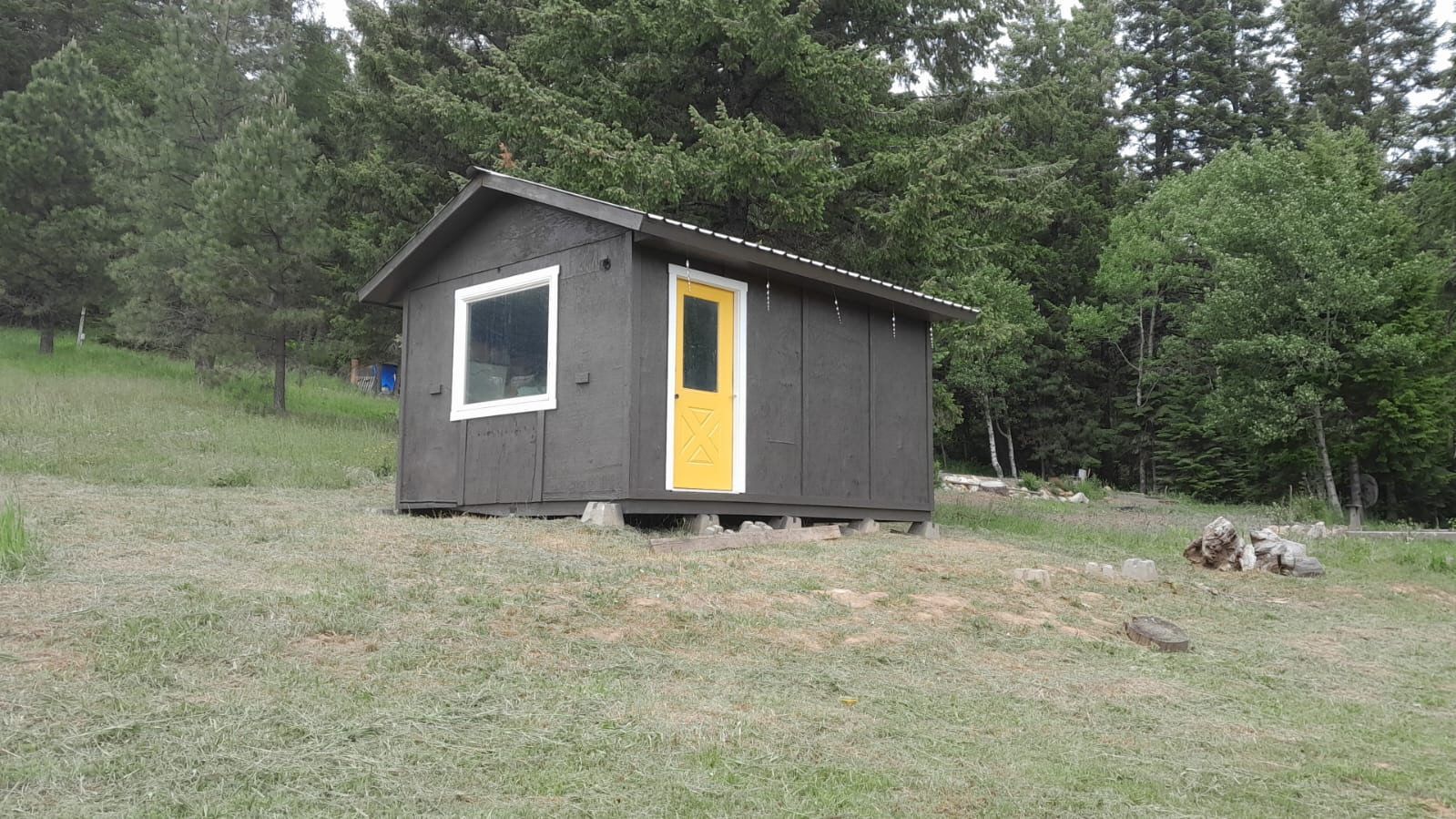 Small dark cabin with a bright yellow door on a grassy hillside, surrounded by trees.