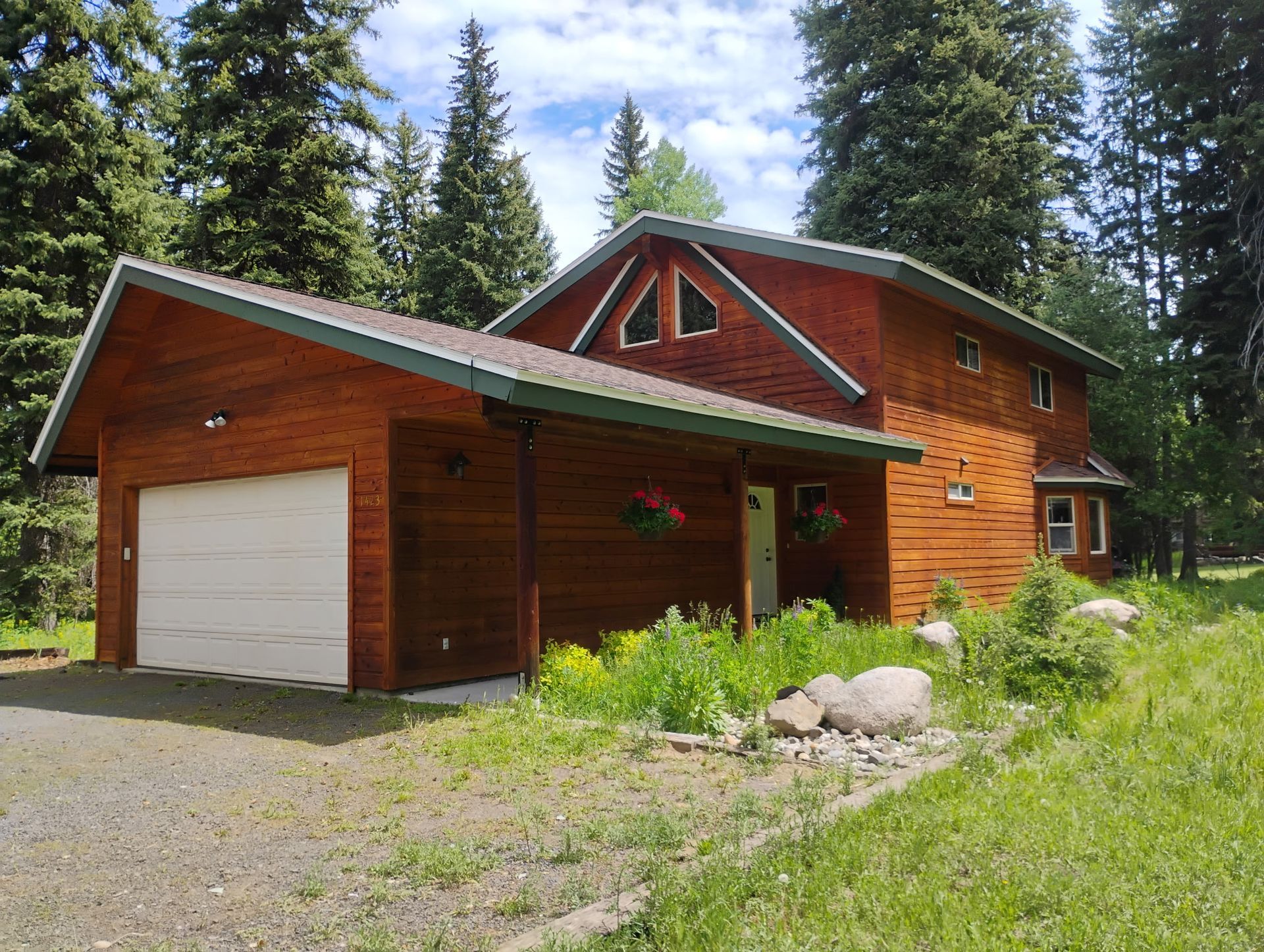 Wooden cabin with attached garage, surrounded by pine trees and greenery under a blue sky.