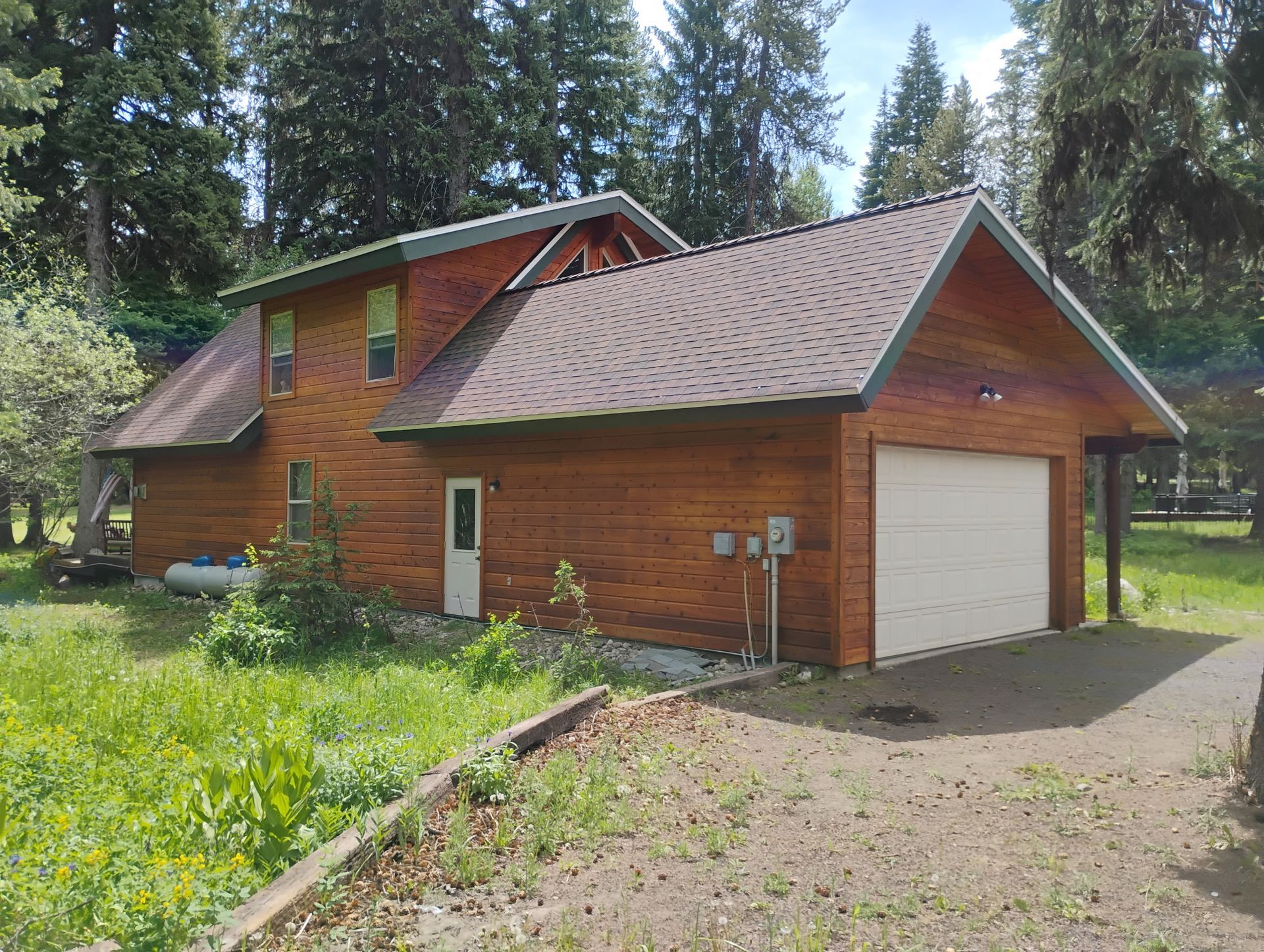 Wooden house with a garage, surrounded by trees and greenery on a sunny day