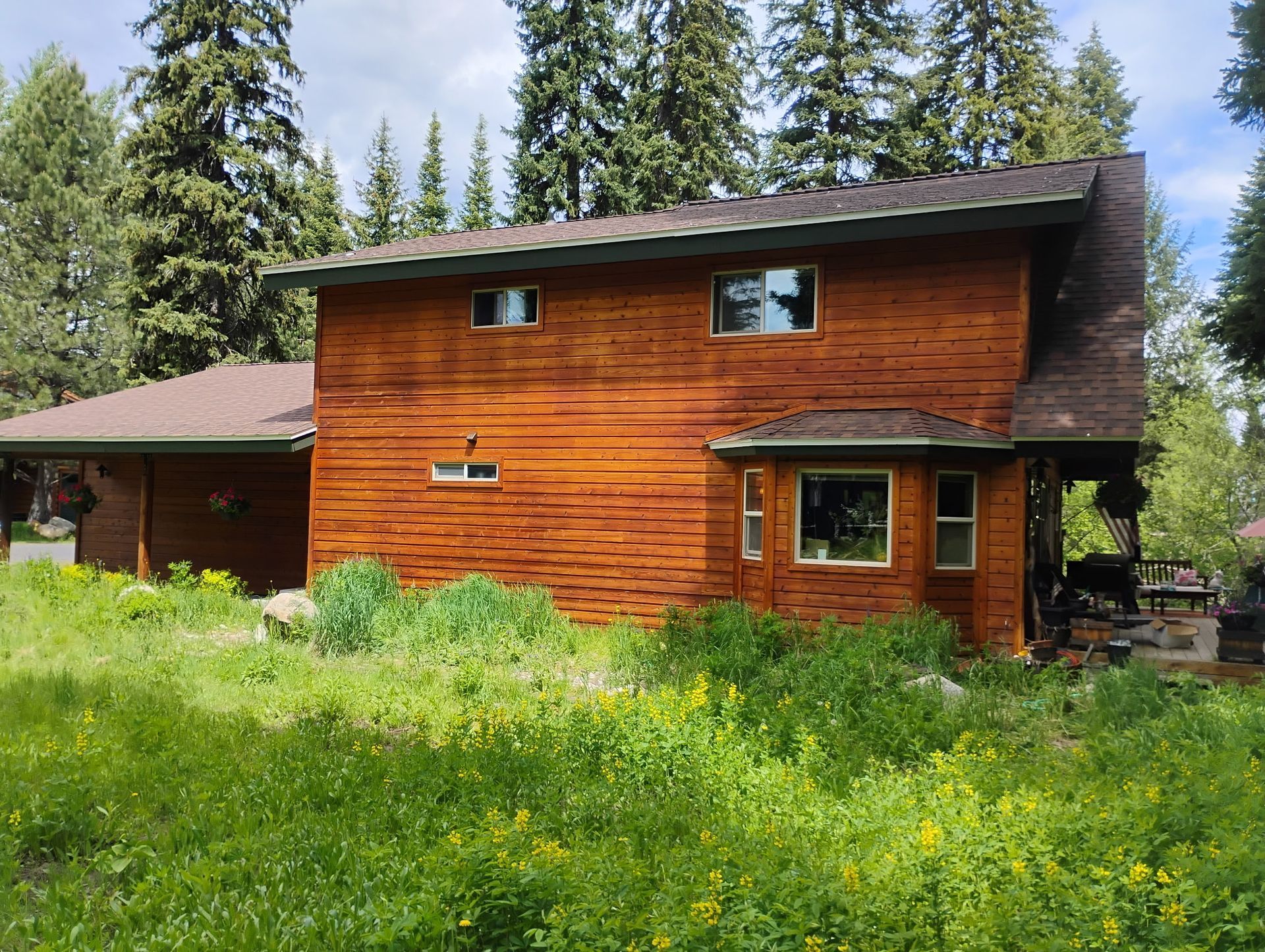Rustic two-story wooden house with green meadow foreground and tall pine trees behind