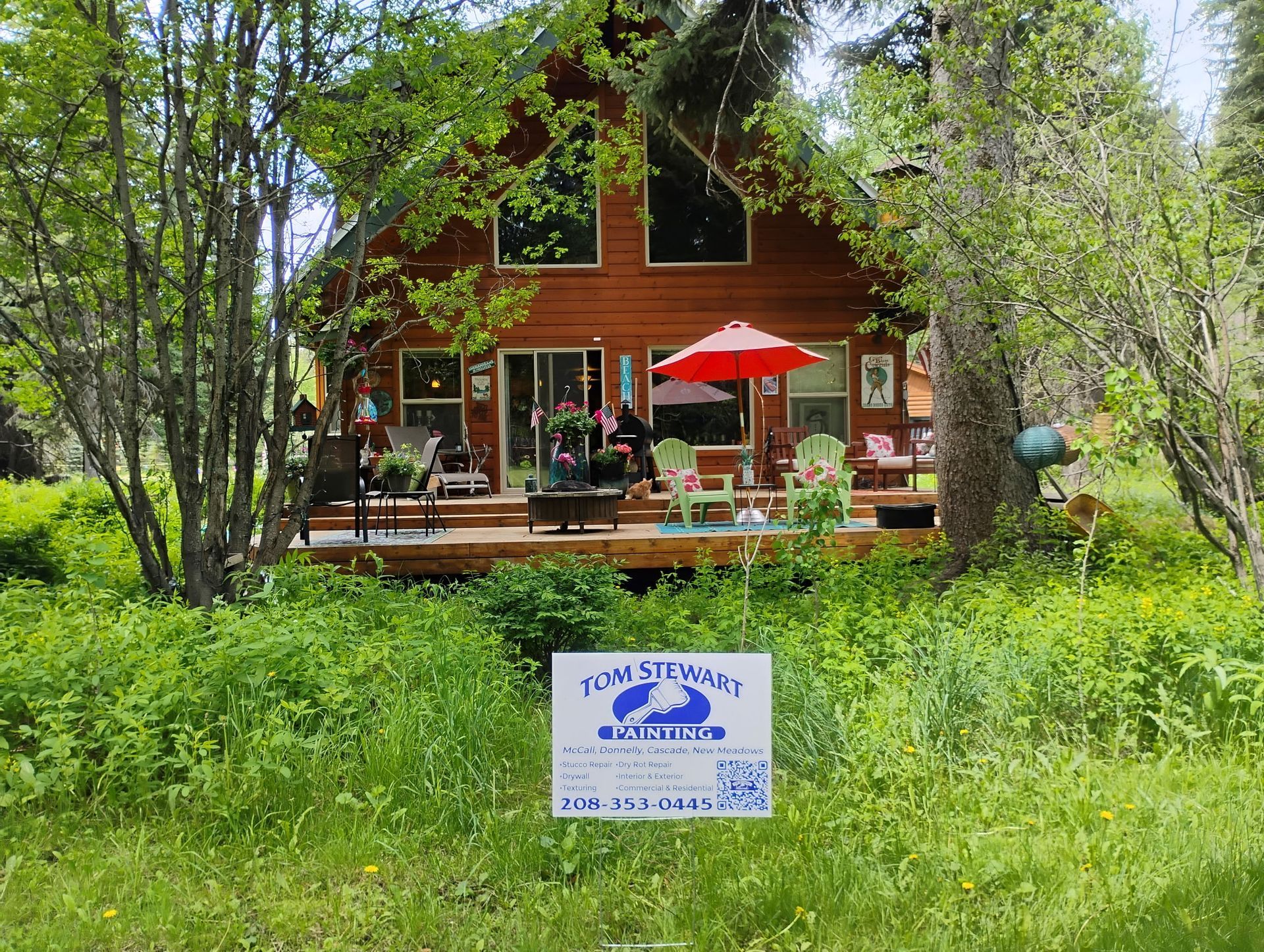 Red cabin among trees with a porch and red umbrella, behind a grassy lawn and a small sign in front