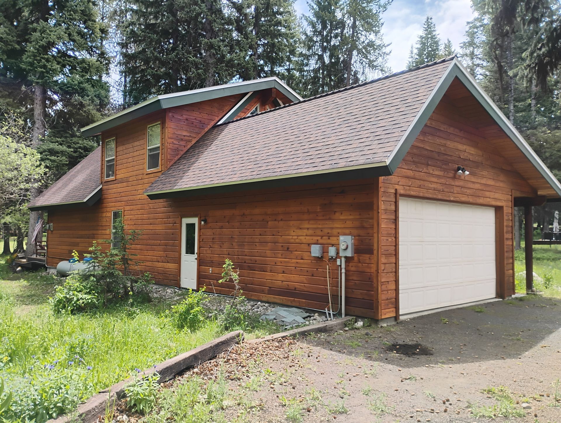 Wooden cabin with garage and green yard surrounded by tall pine trees