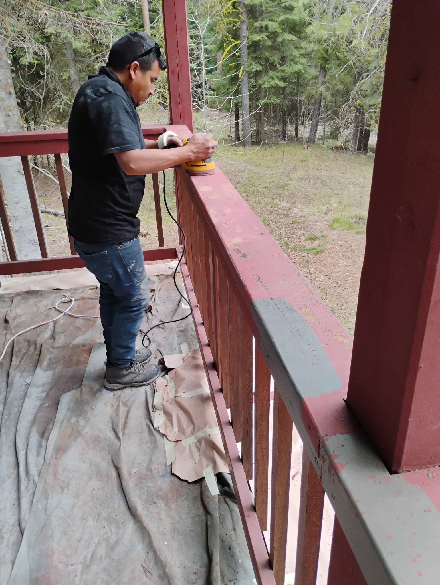 Man standing on a porch railing, looking out over a wooded yard and holding a cup.