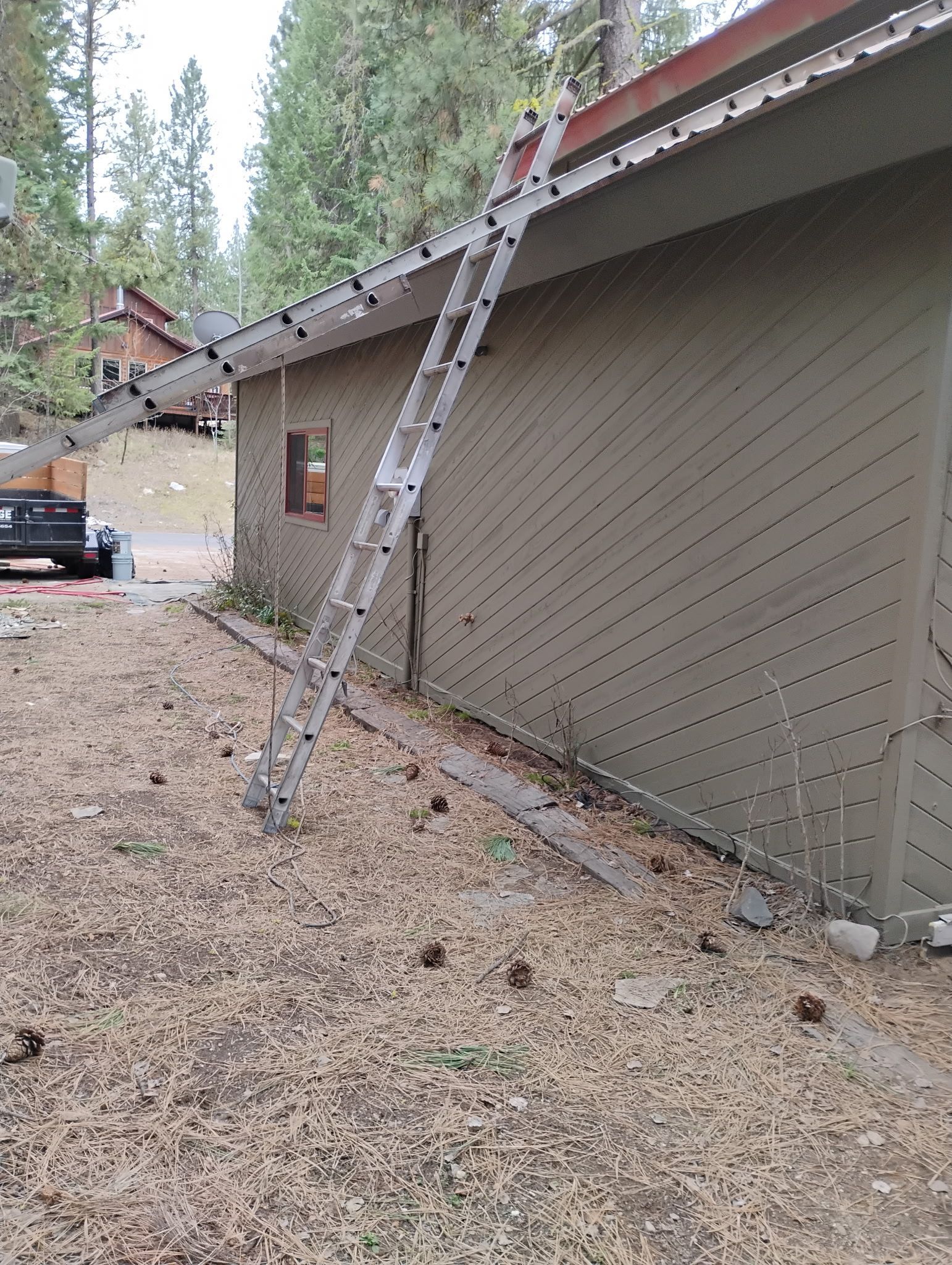 Leaning ladder against tan house wall with debris on gravel ground near a pine forest