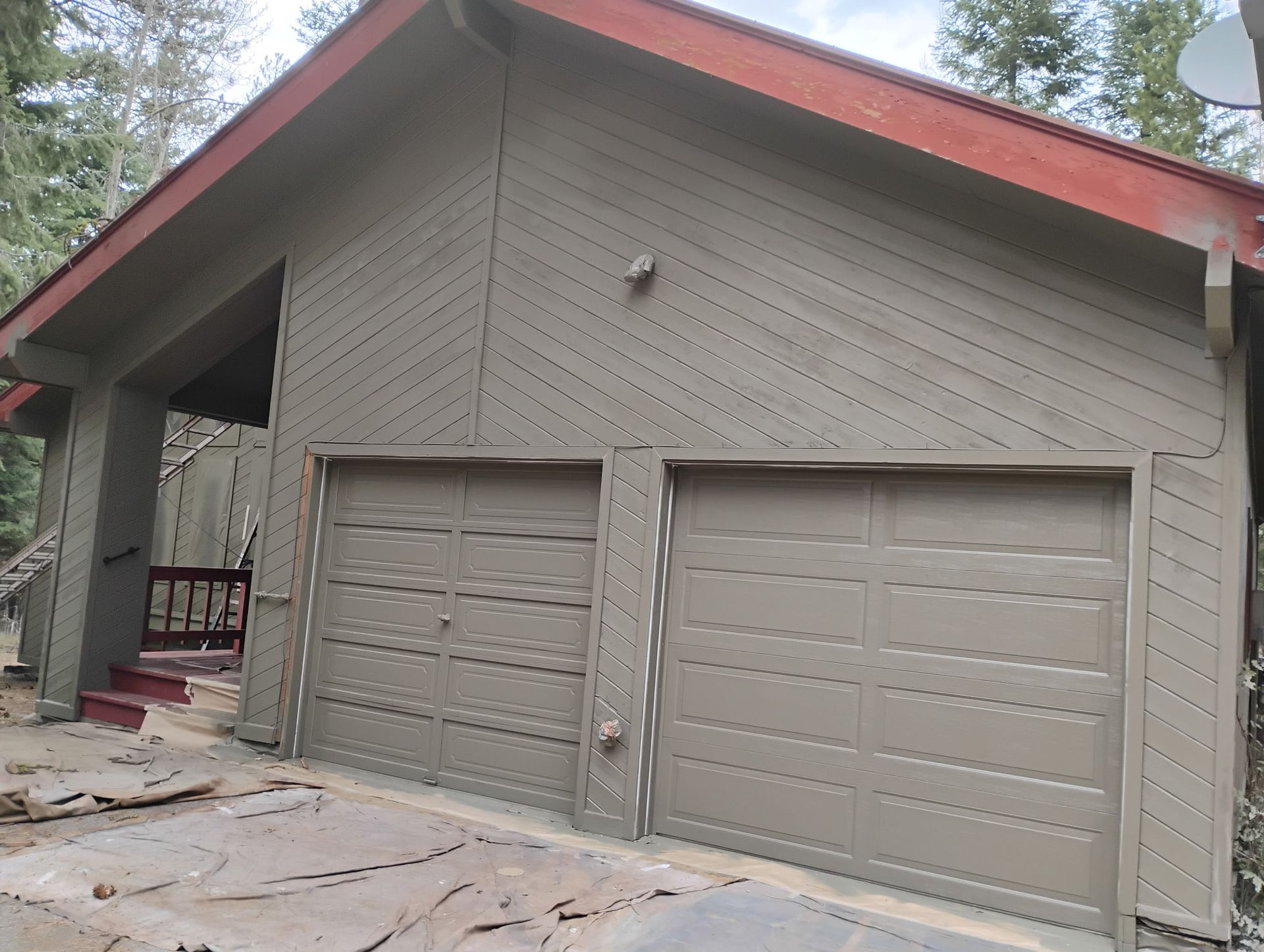 Gray two-car garage with red roof trim and a small covered porch in a wooded setting