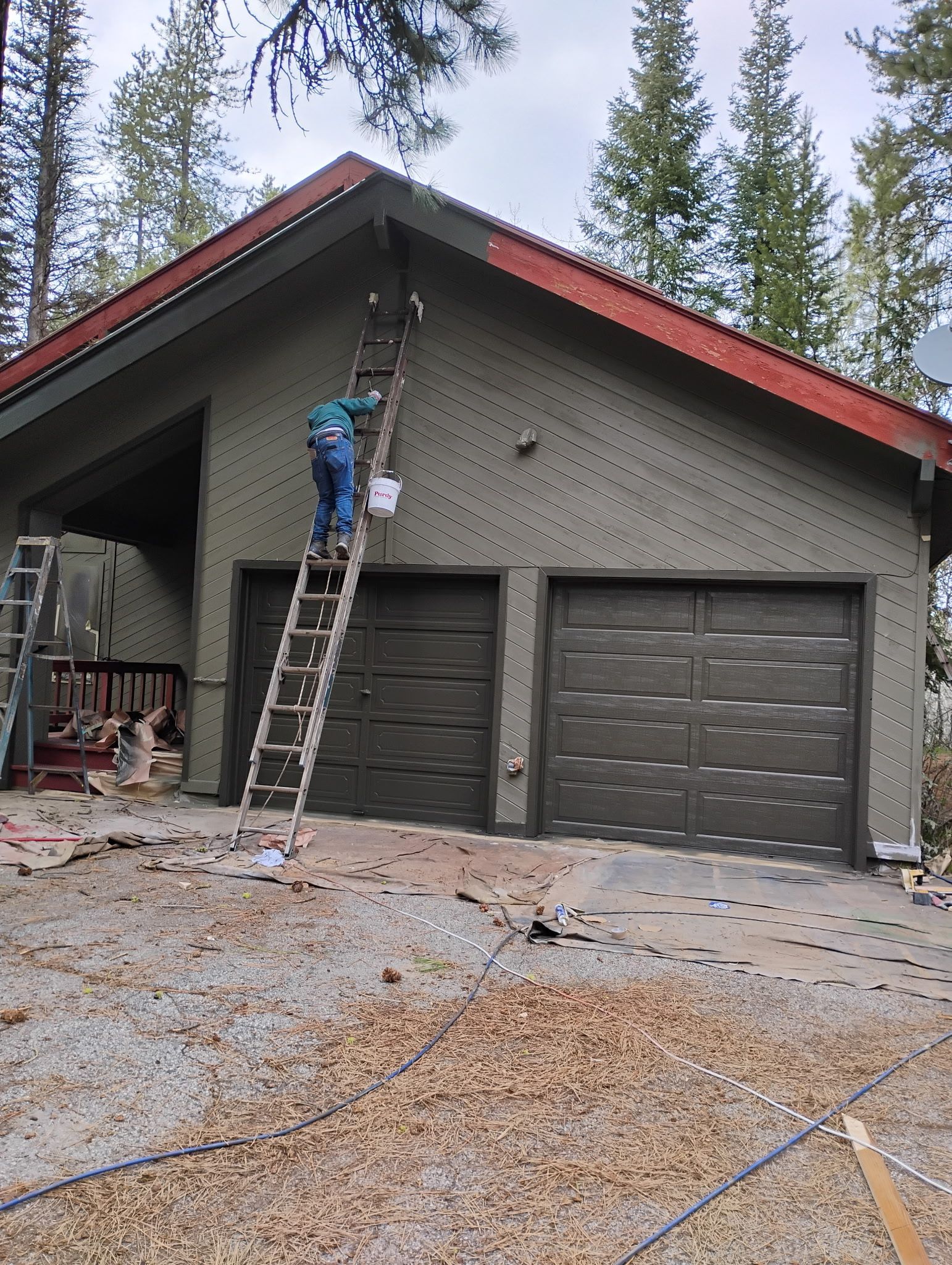 Person on ladder cleaning the gable of a gray garage with red trim and two closed doors