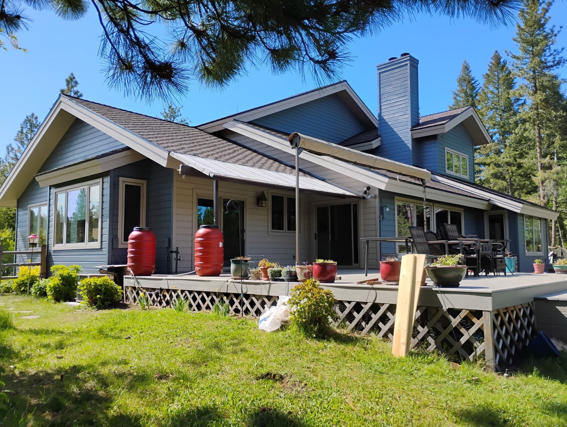 Blue lakeside house with wraparound porch, red propane tanks, and green lawn under tall trees.