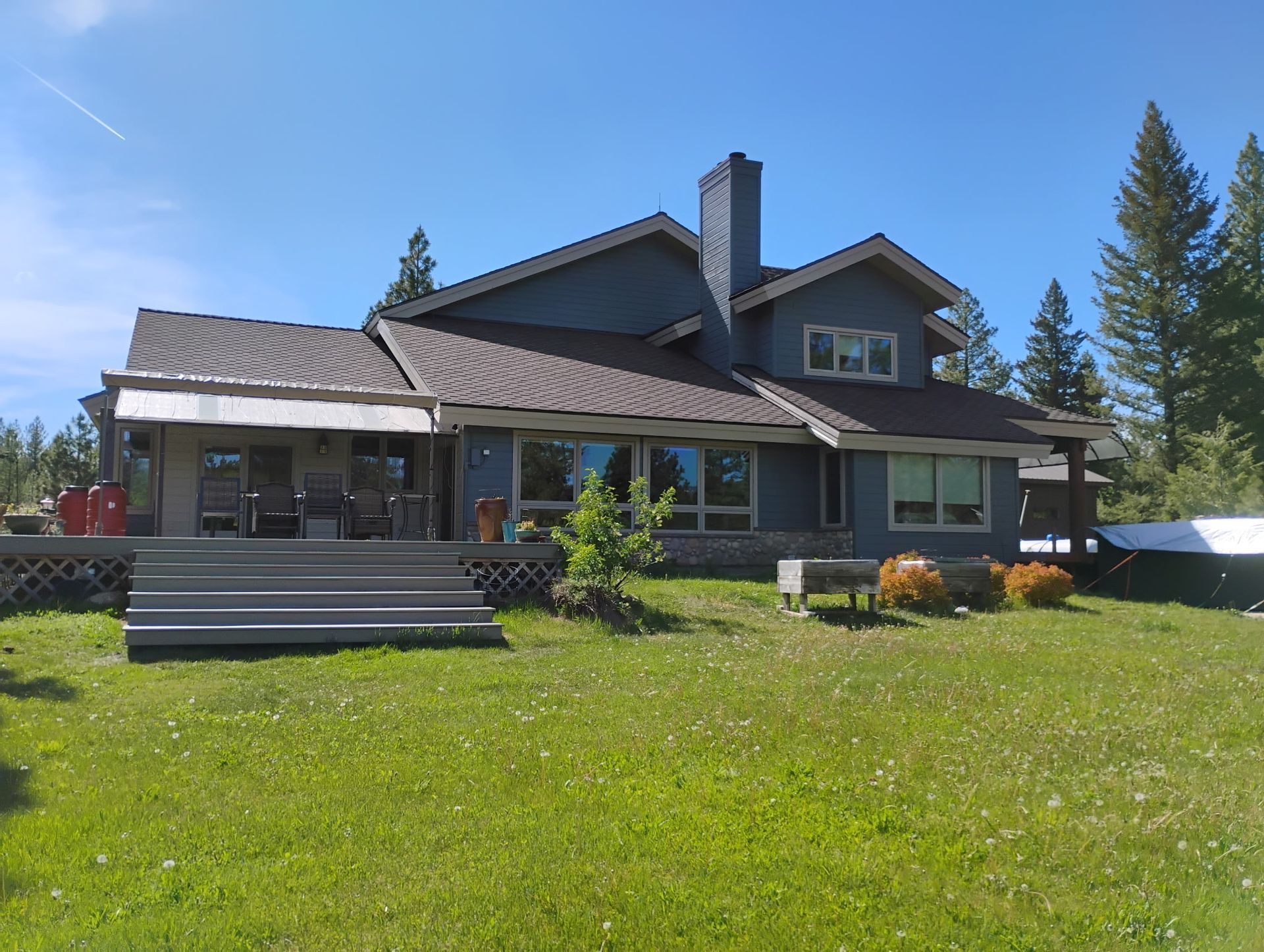 Large rustic house with a covered porch, green lawn, and tall pine trees under a blue sky