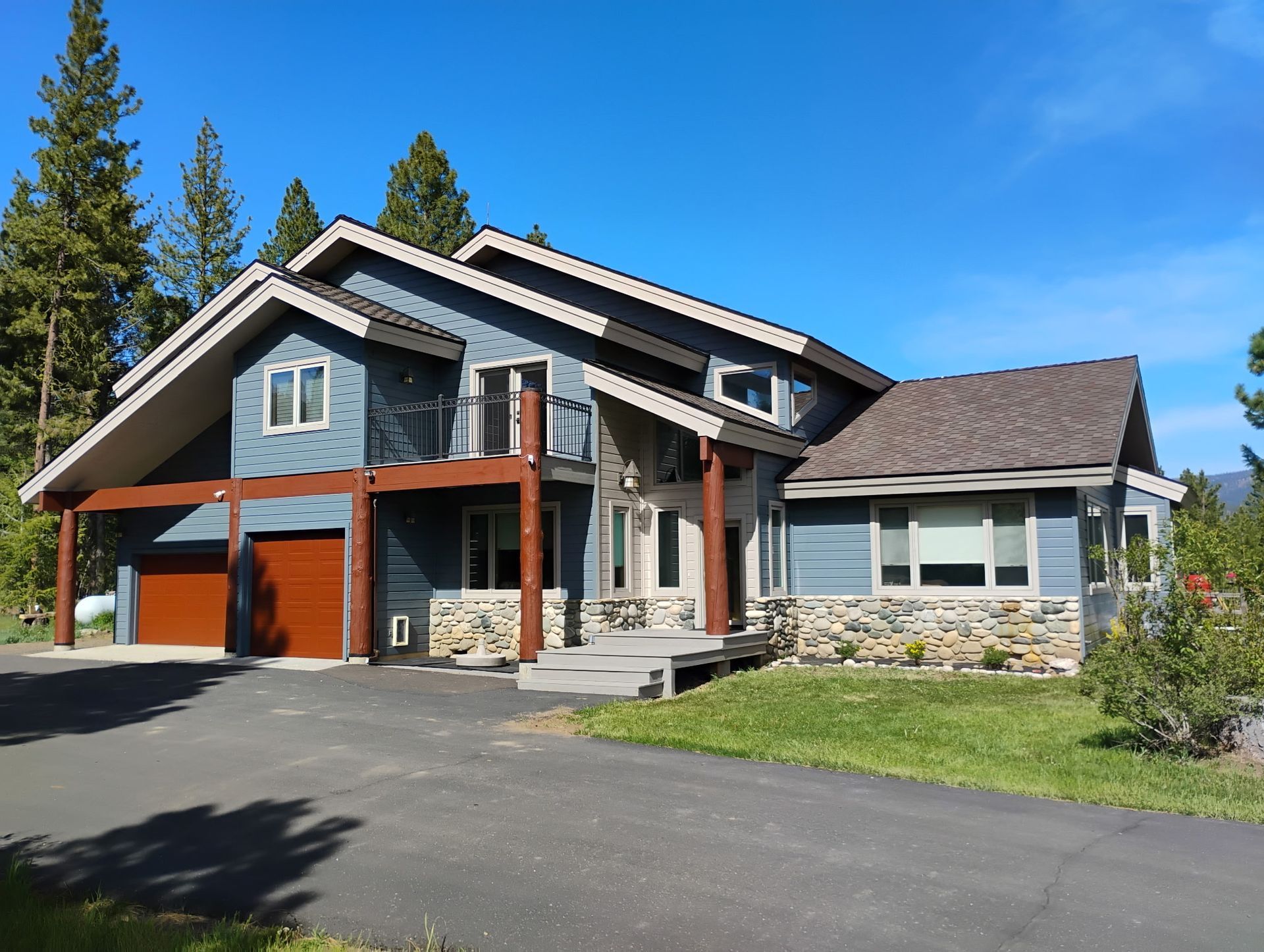 Modern two-story house with stone and wood exterior, front porch, garage, and paved driveway under a blue sky