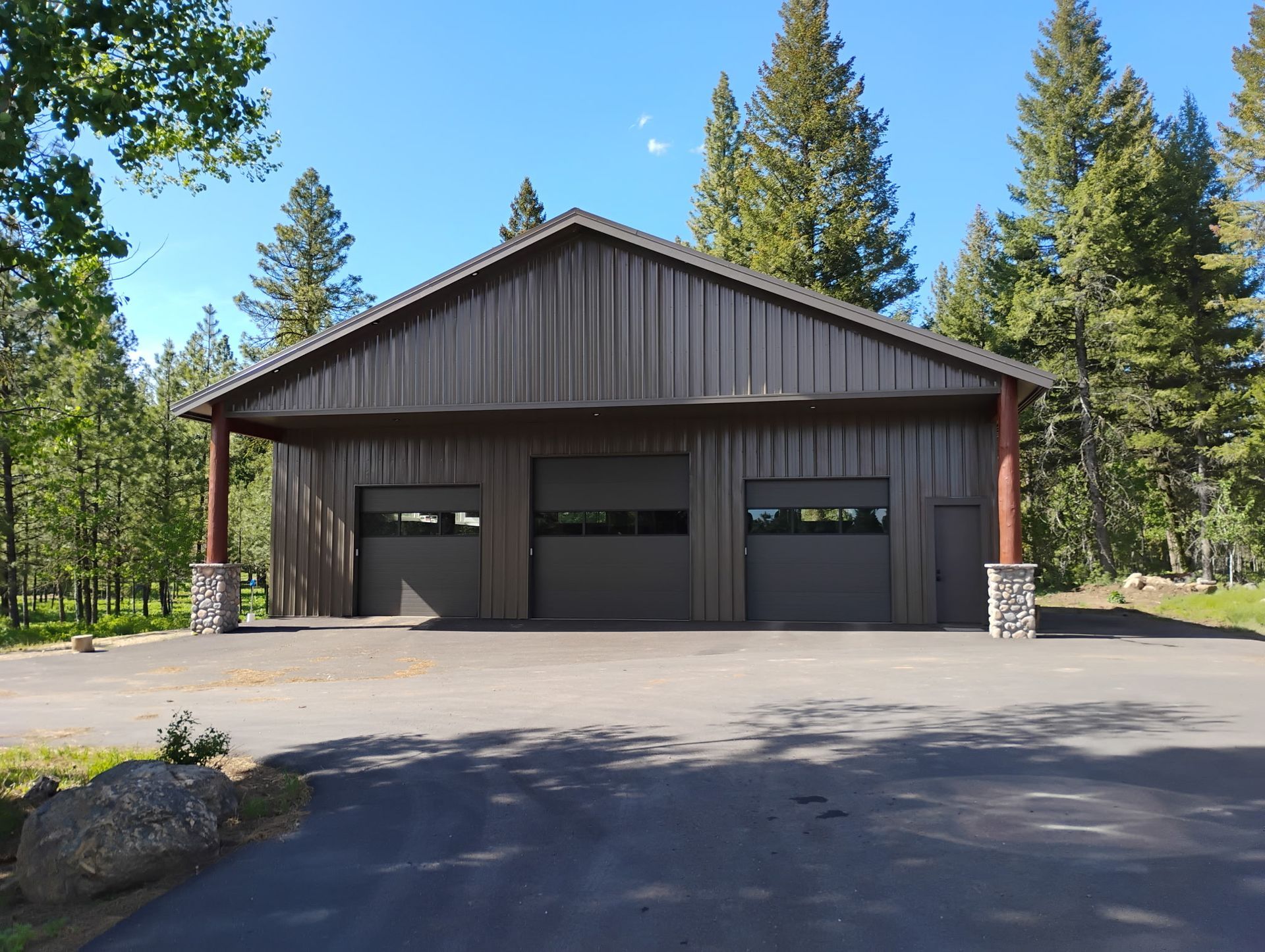 Rustic wooden garage with three dark doors, set among tall pine trees under a clear blue sky