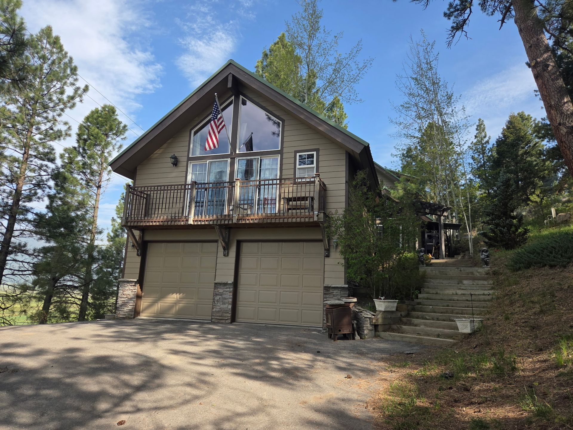 A small cabin with a balcony and garage under tall trees on a sunny hillside.