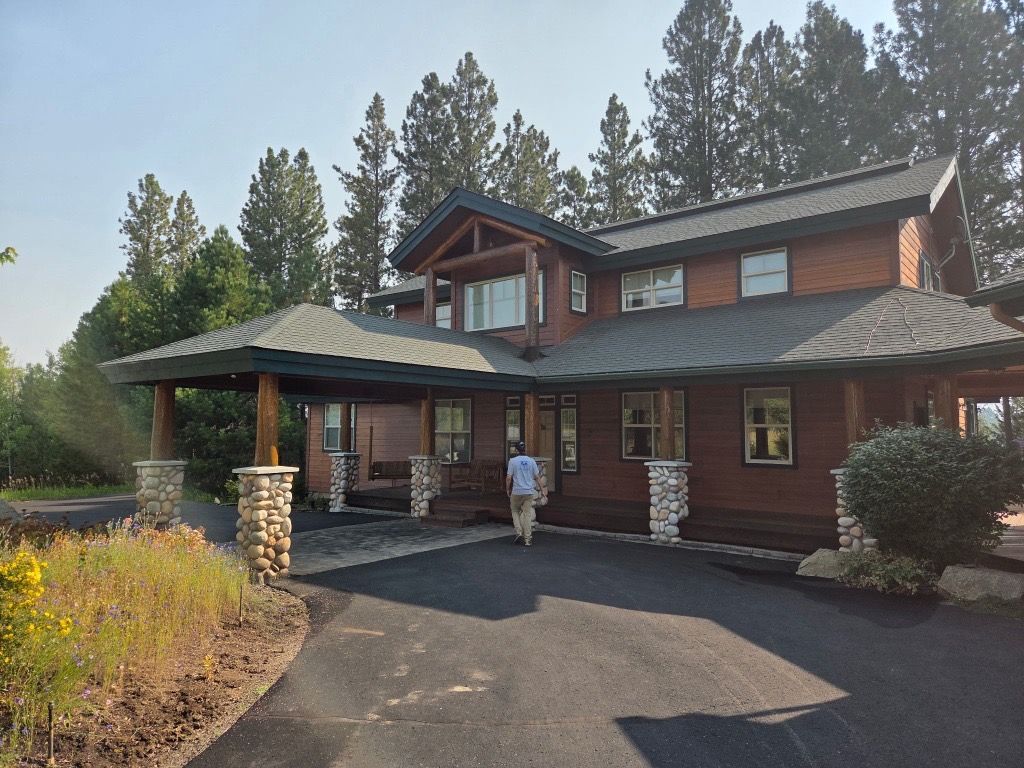 Wooden lodge-style house with covered entrance, stone pillars, and paved driveway surrounded by trees.