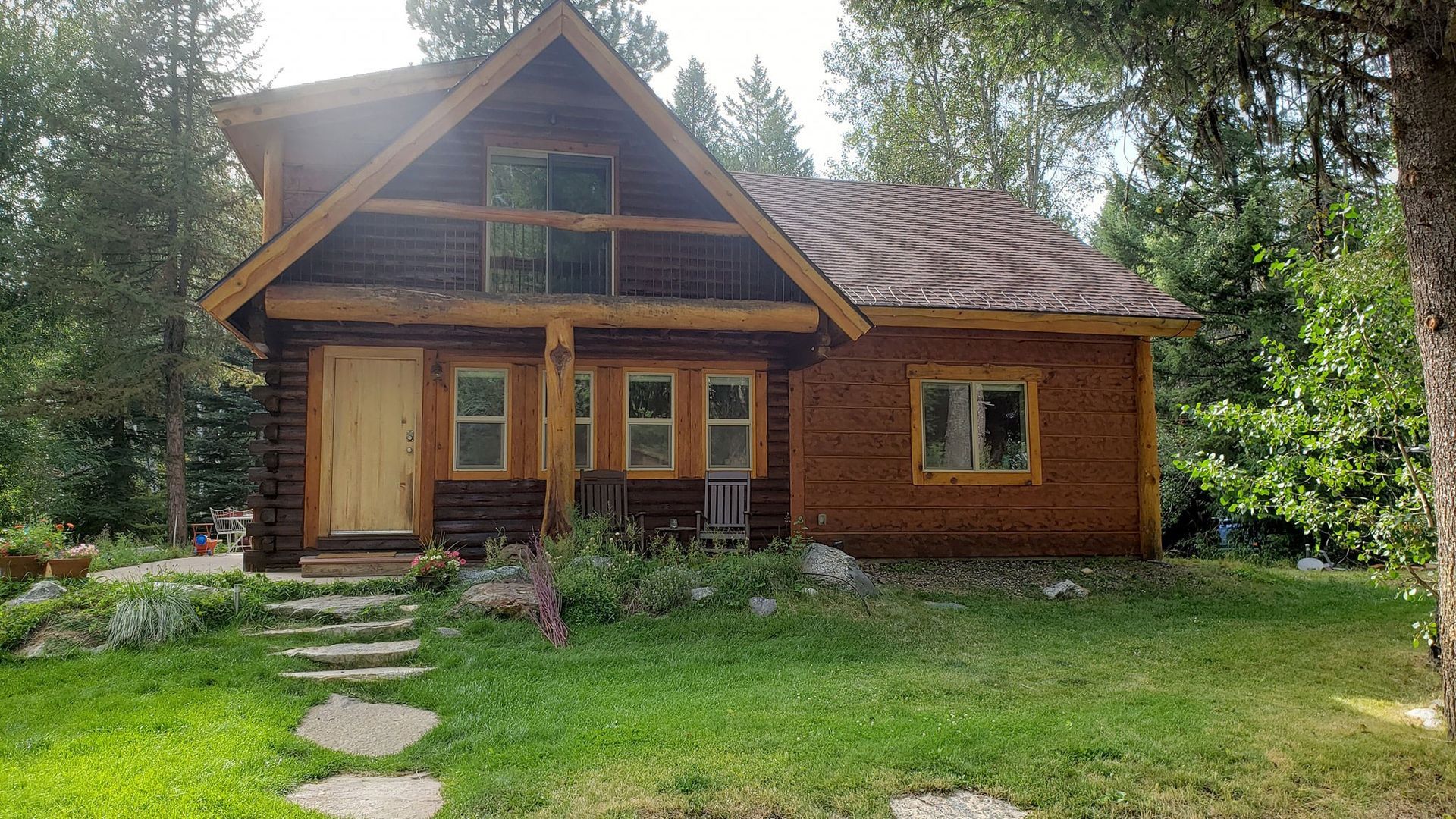 Wooden cabin with a steep roof and stone path, surrounded by trees and green lawn.