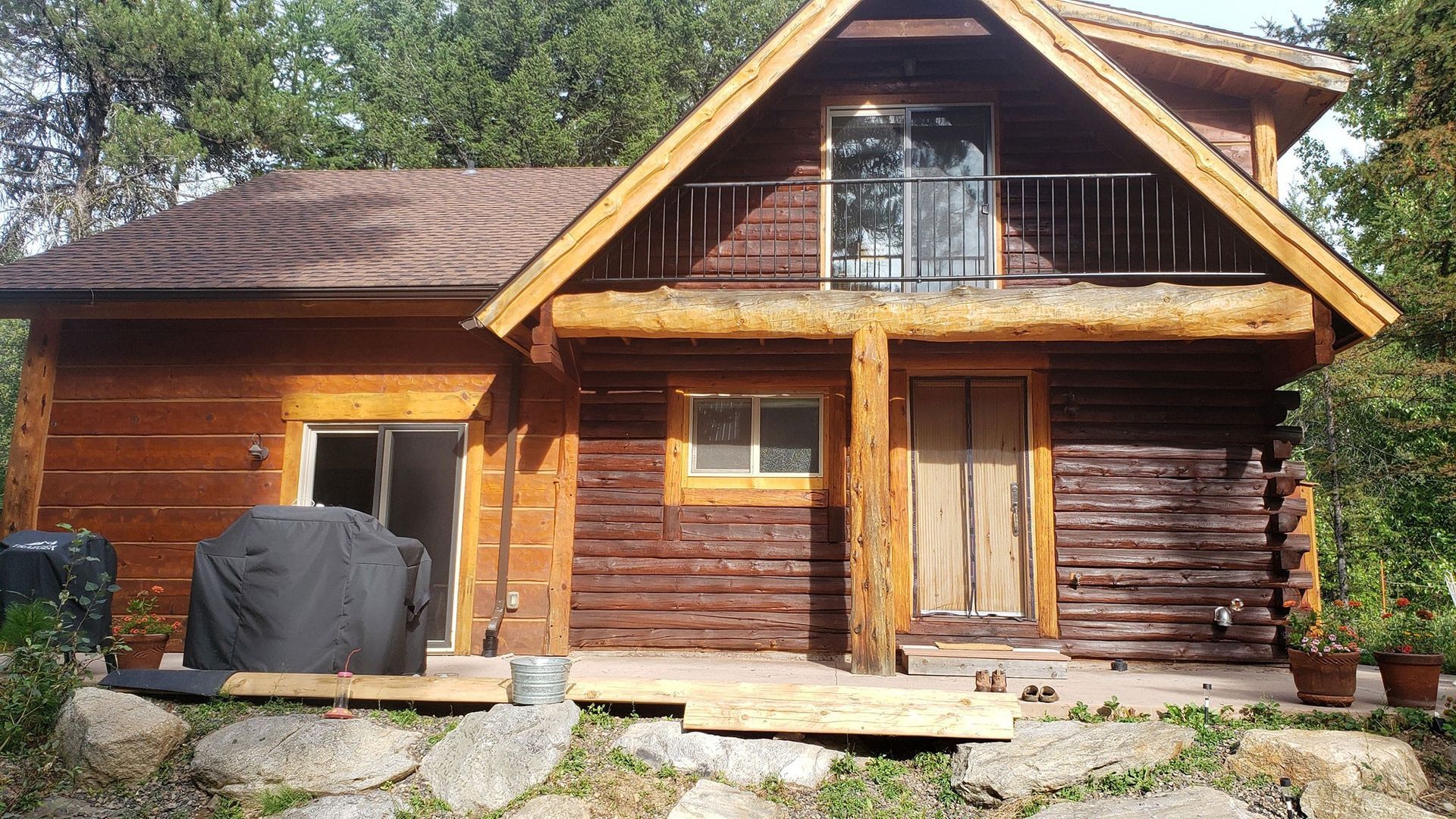 Rustic wooden cabin with a porch, stone steps, and a black grill in front, surrounded by trees