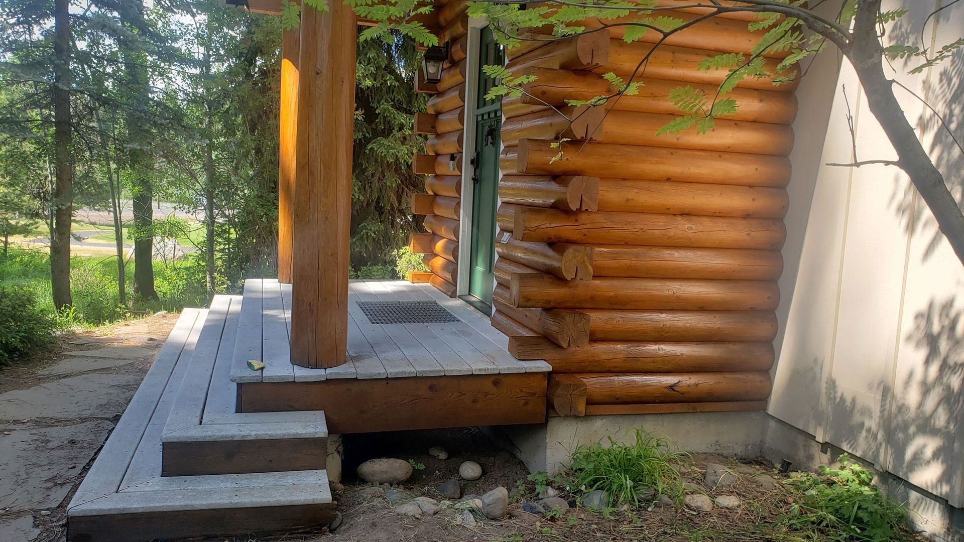 Wooden porch with concrete steps beside a log cabin wall, surrounded by trees and greenery.