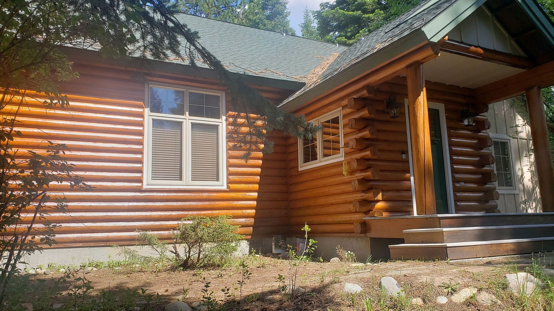 Log cabin exterior with a covered porch, white-framed window, and steps in a sunny wooded setting