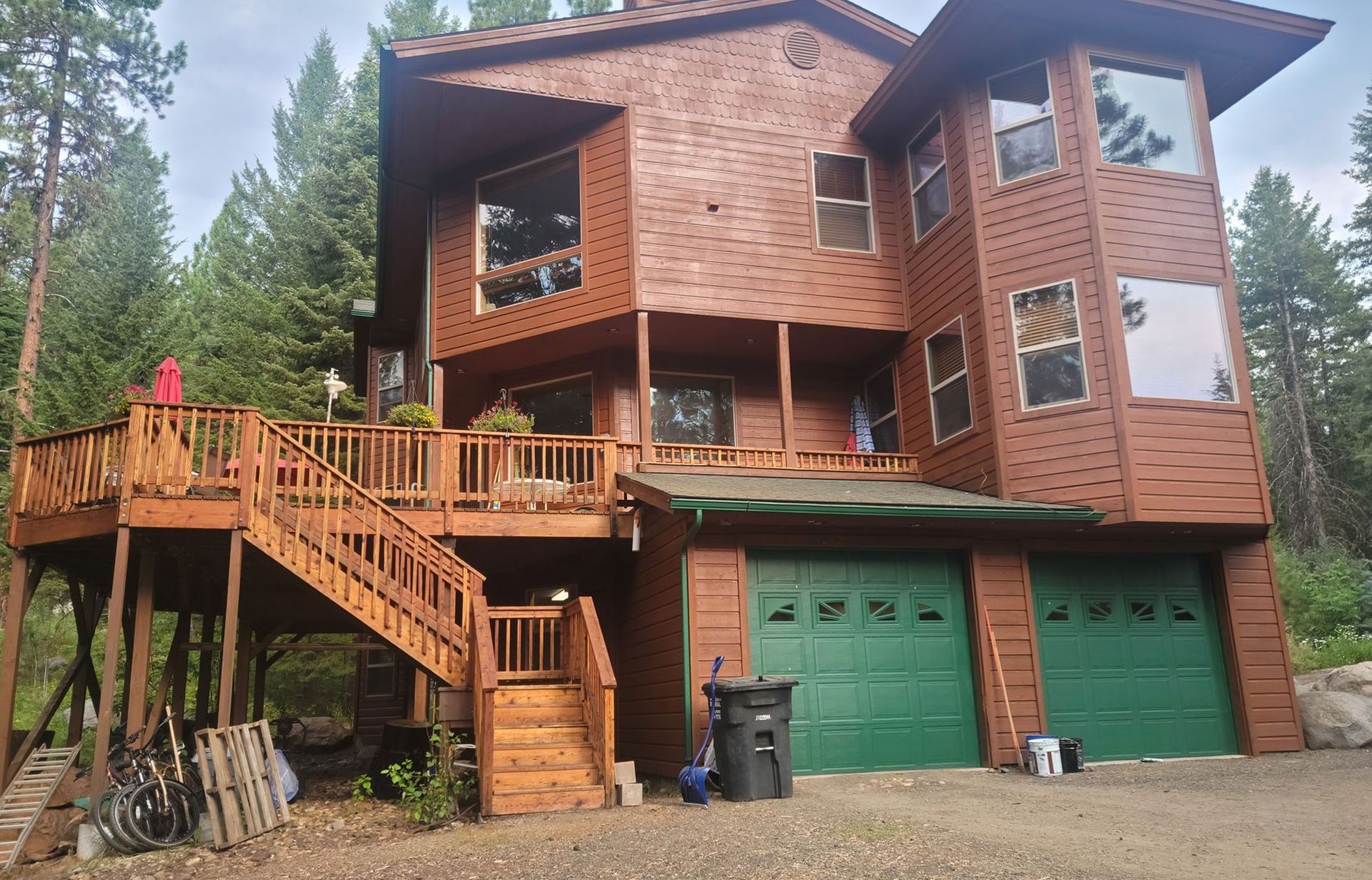 Wooden two-story house with large deck, green garage doors, and stairs in a wooded setting