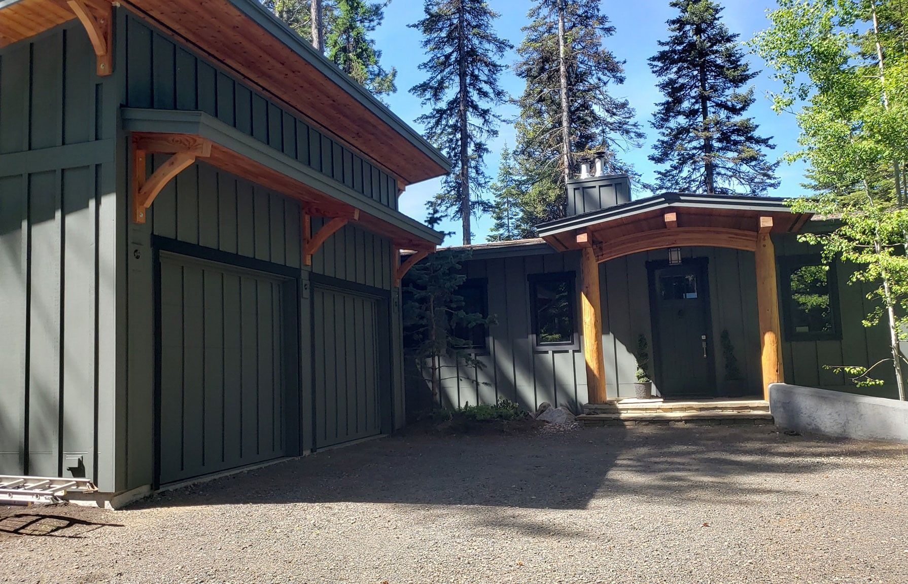 Modern dark-green and tan house exteriors with gravel driveway and tall pine trees in bright sunlight