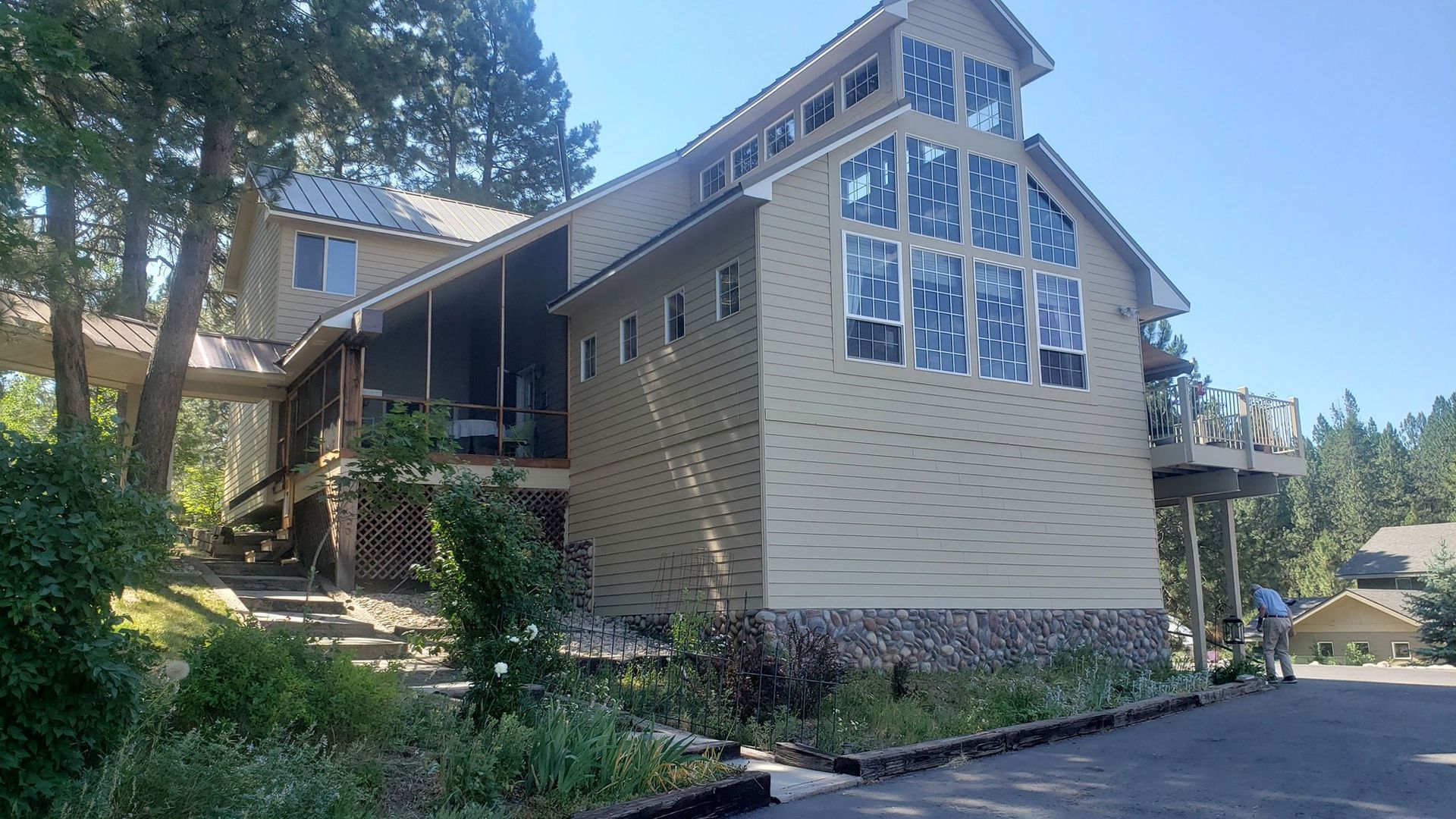 Large beige house with tall windows and a sloped roof among trees on a sunny hillside