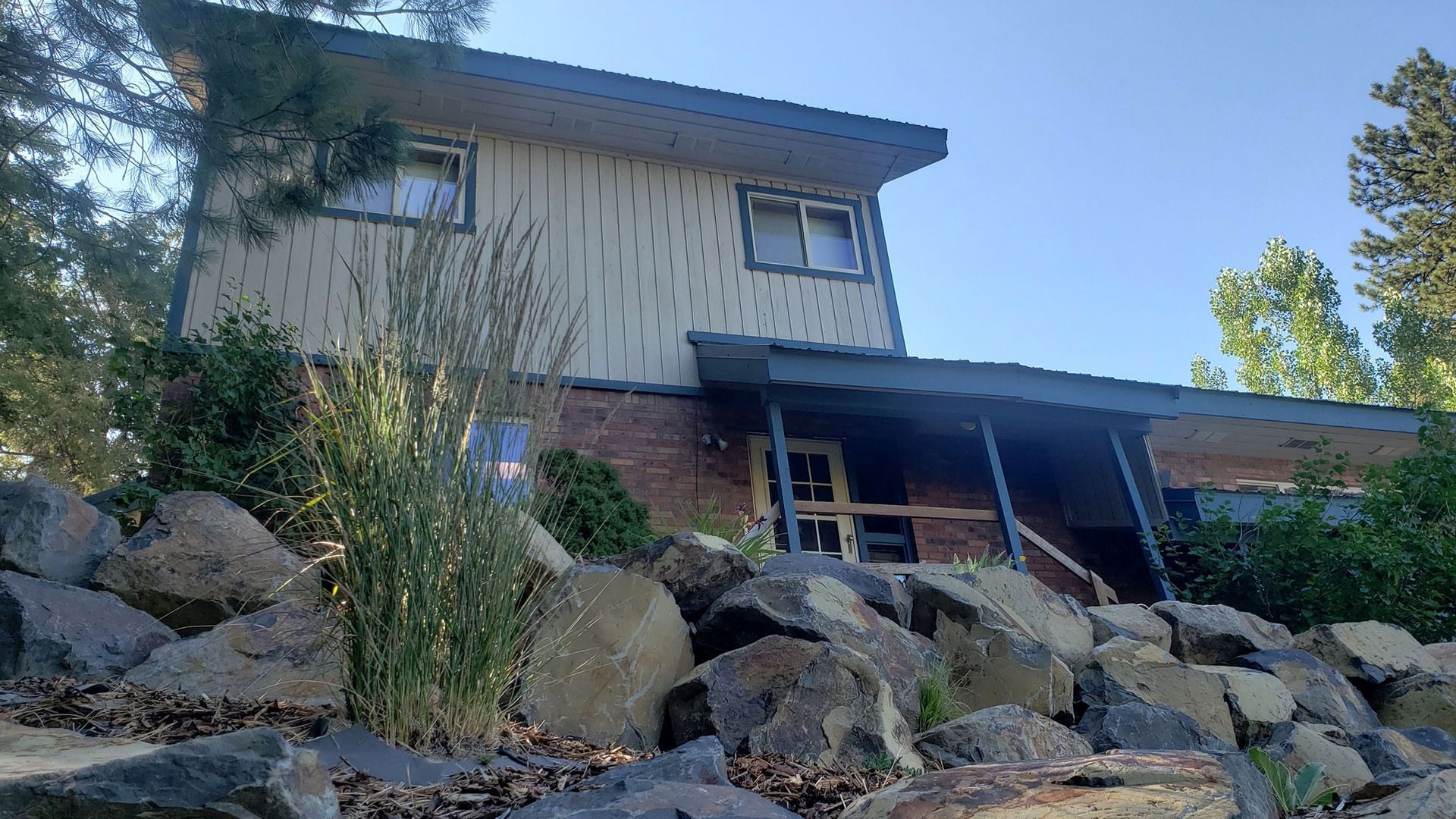 Two-story house with a covered porch on a rocky hillside under a clear blue sky