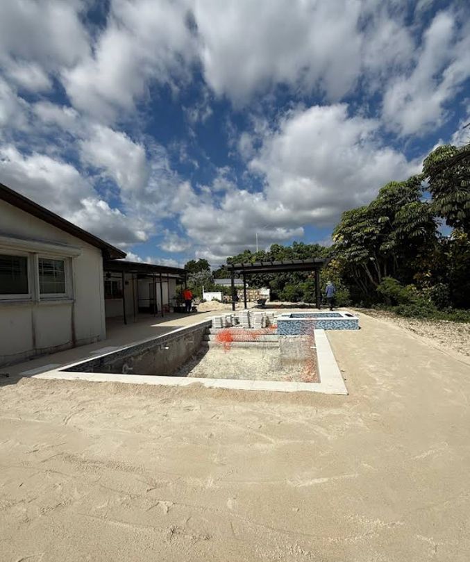 Outdoor courtyard with a long narrow pool, white buildings, sandy ground, and a cloudy blue sky.