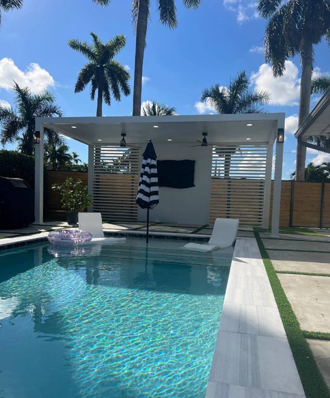 Backyard pool with turquoise water, lounge chairs, striped umbrella, and palm trees under a blue sky