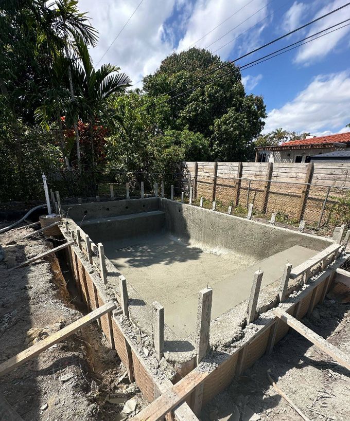 Concrete pool foundation under construction in a backyard, with rebar stakes and wooden formwork.