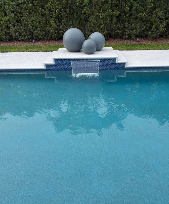 Blue swimming pool with three gray decorative spheres on the deck against a hedge backdrop