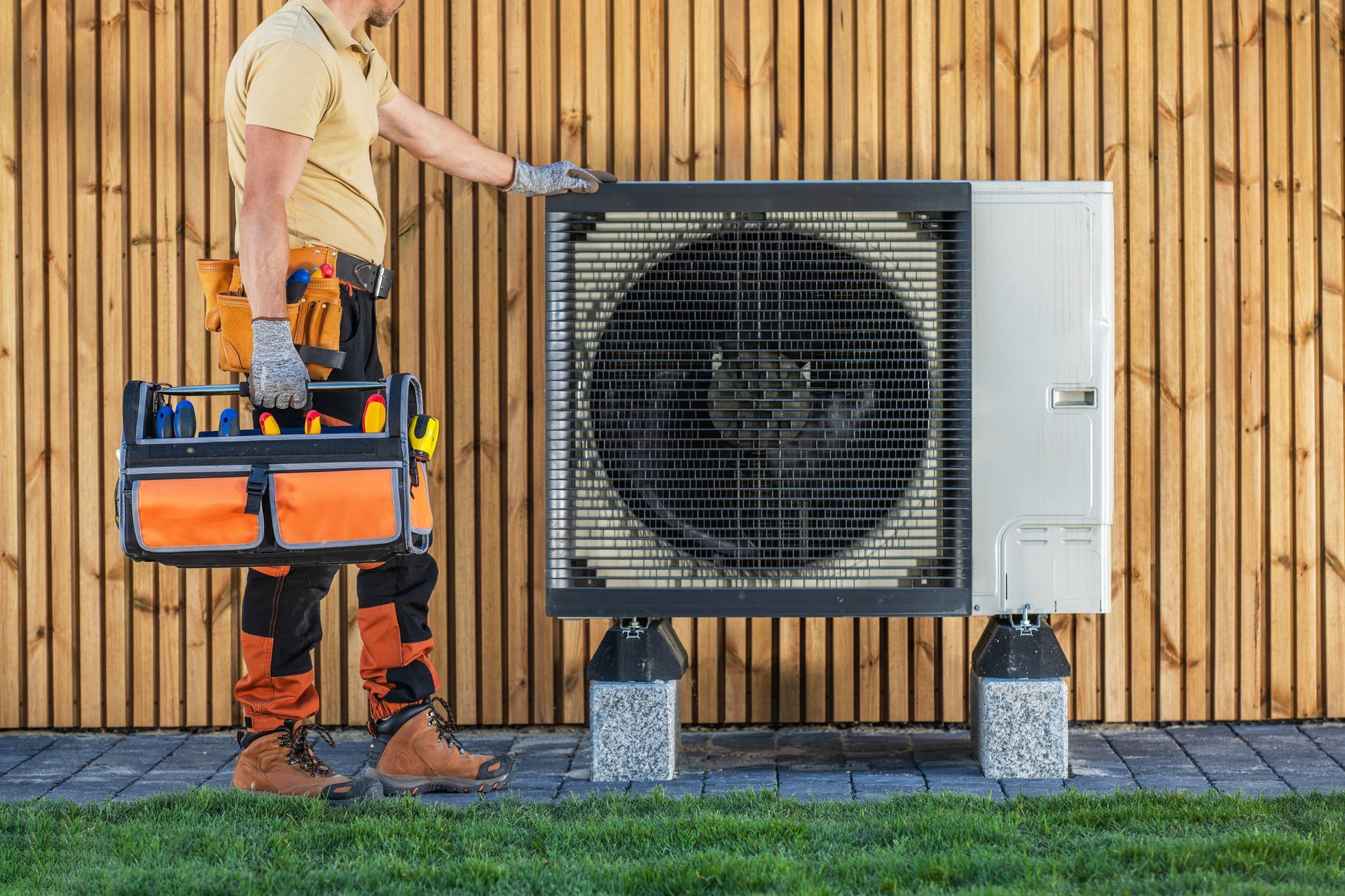 A technician holding a toolbox inspects an outdoor heat pump unit, standing on a brick base, against a wooden wall.