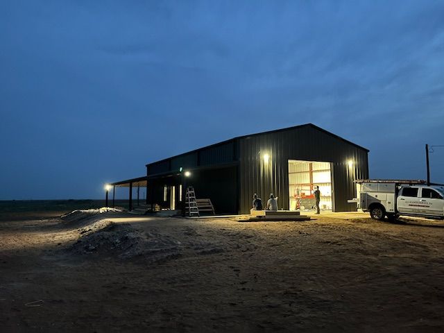 A dark metal building at dusk with exterior lights on. A work truck is parked nearby.