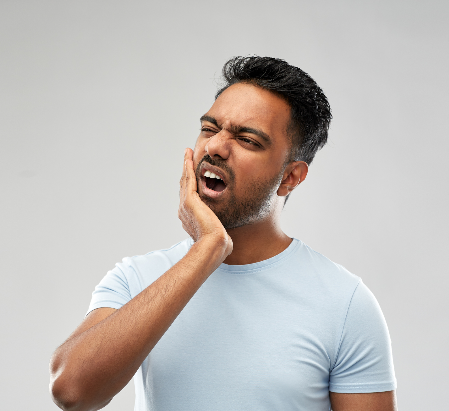 Man in a light blue shirt clutching his cheek in pain against a plain gray background