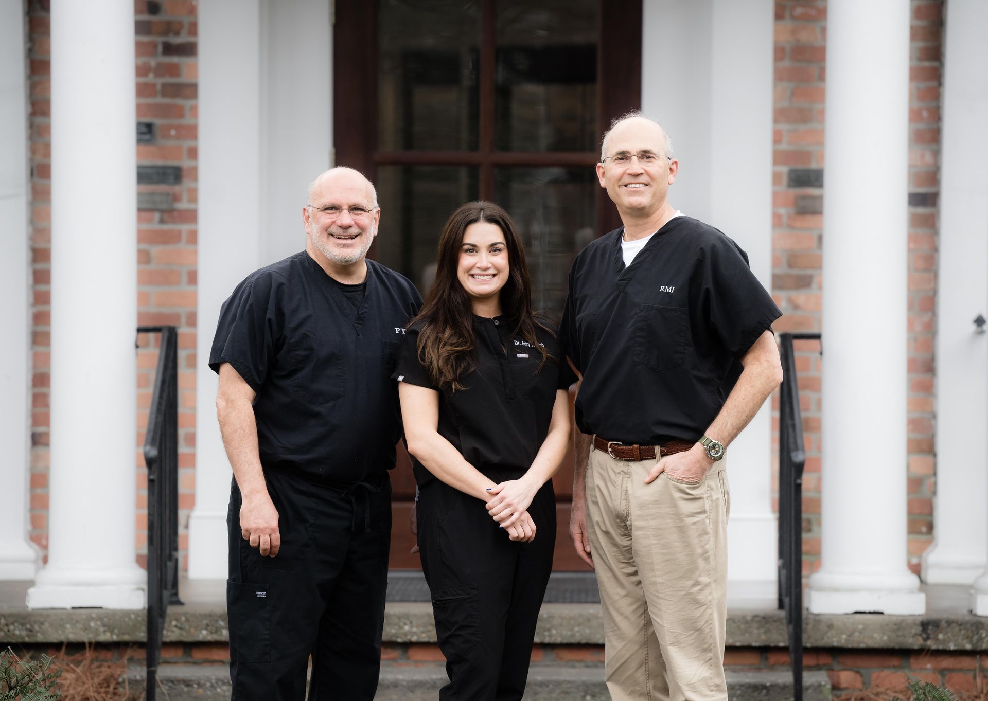 Three people in black tops standing in front of a building entrance with columns and brick walls