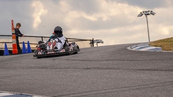 Go-kart racing on track. Driver in helmet turns corner, with cones and spotter present. Cloudy sky in background.