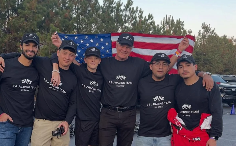 Group of six people in matching black shirts pose with an American flag; outside.
