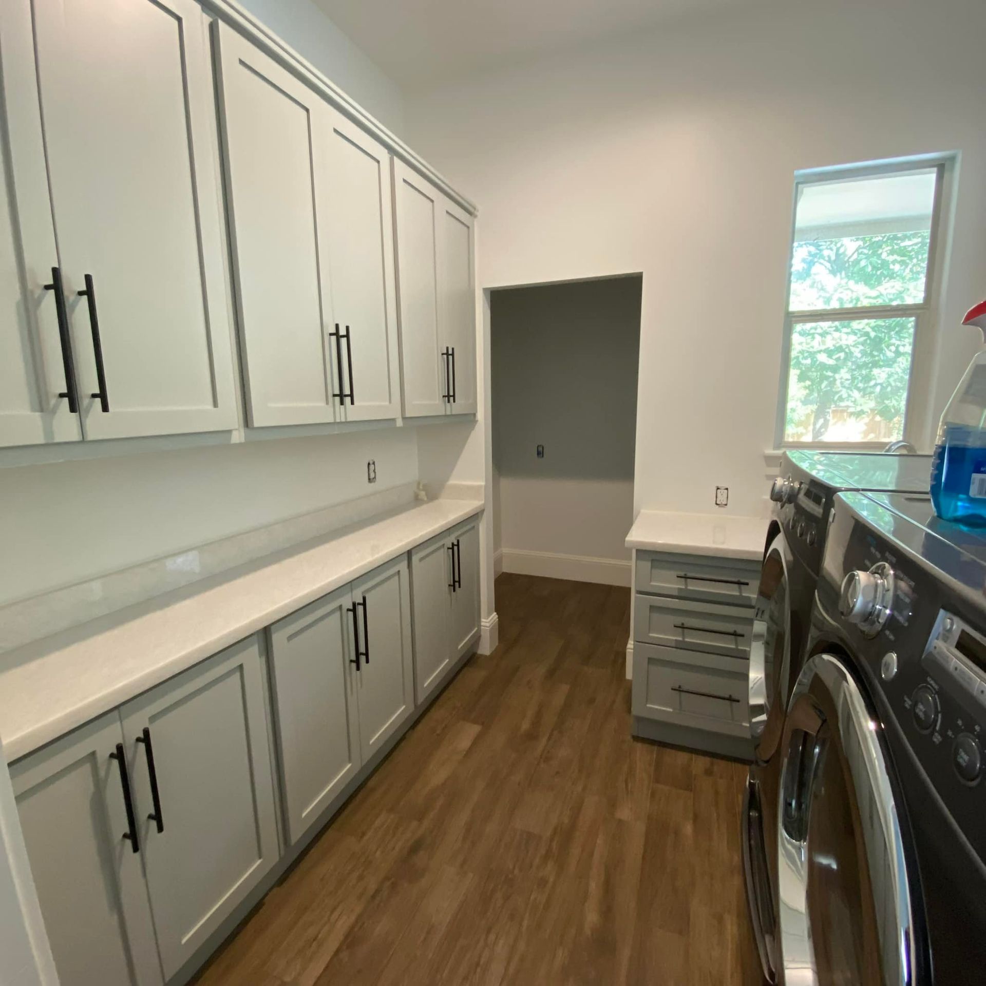 A laundry room with white cabinets, light-colored countertops, wood-look flooring, and a dark front-loading washer.