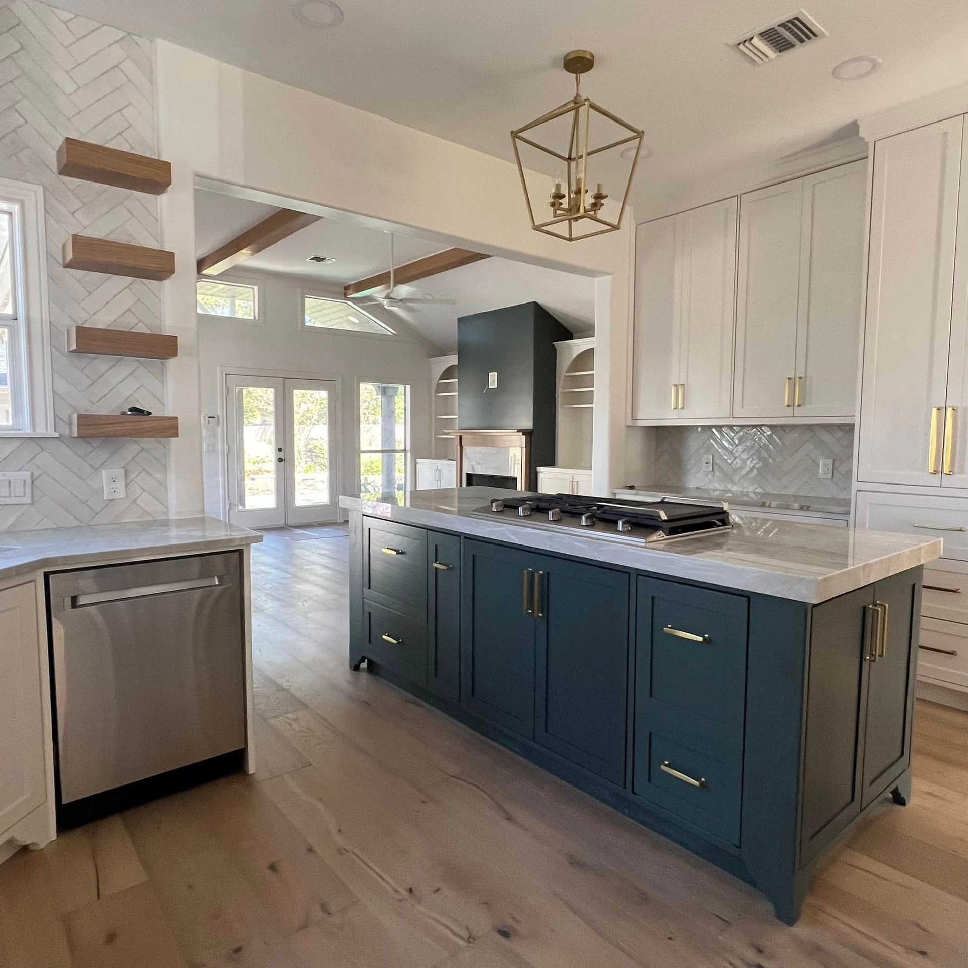 Modern kitchen with dark teal island, marble countertops, white cabinets, and floating wood shelves against herringbone tile.