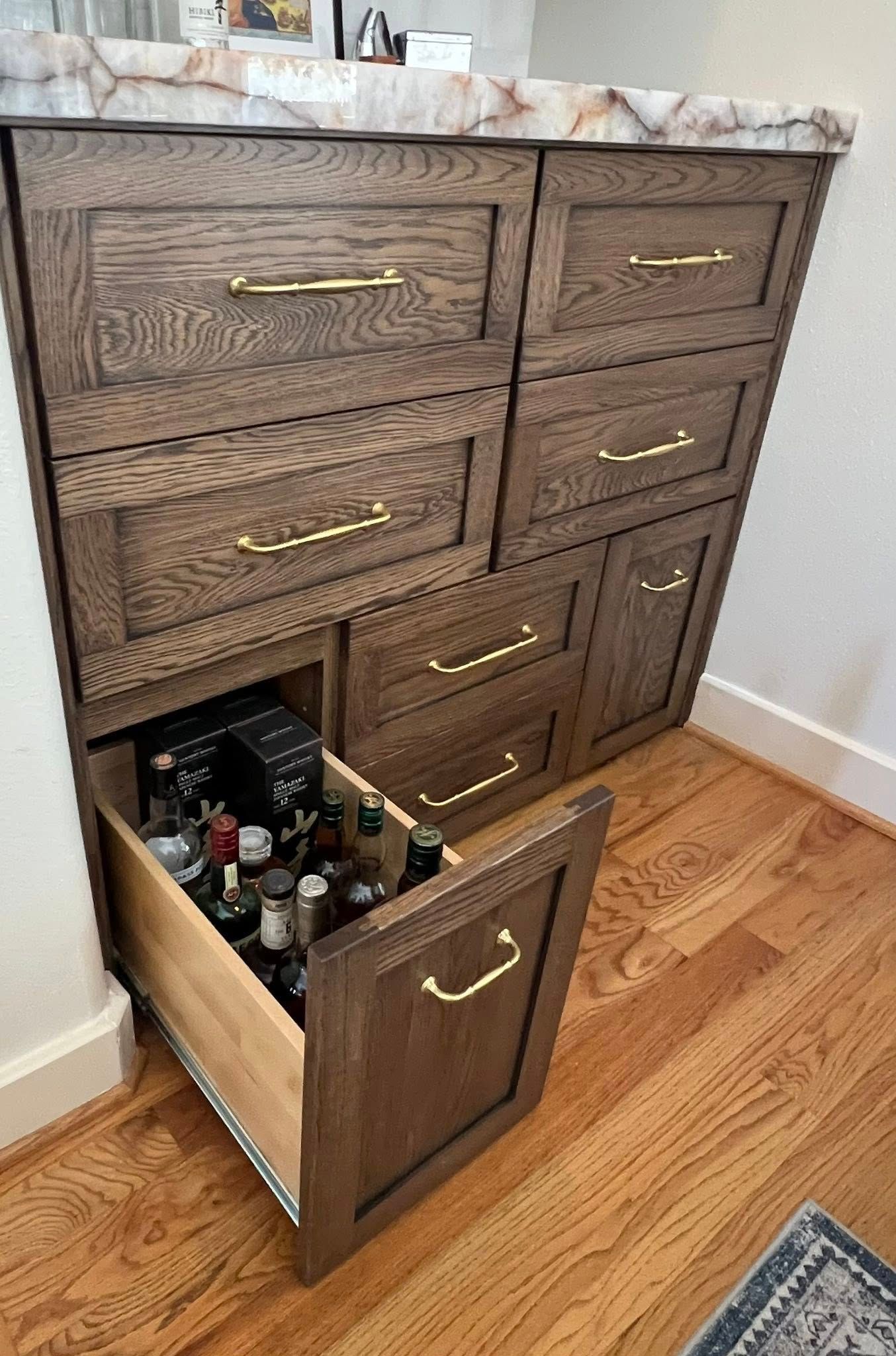 A dark wood cabinet with brass hardware, featuring one open bottom drawer filled with glass liquor bottles.
