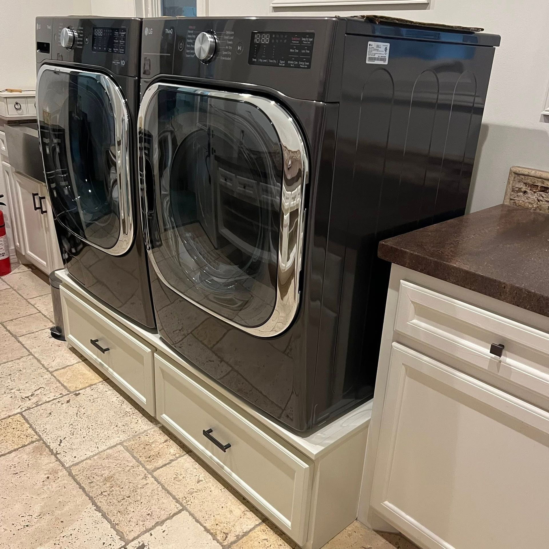 Two dark gray washing machines sitting on white storage pedestals in a tiled laundry room.