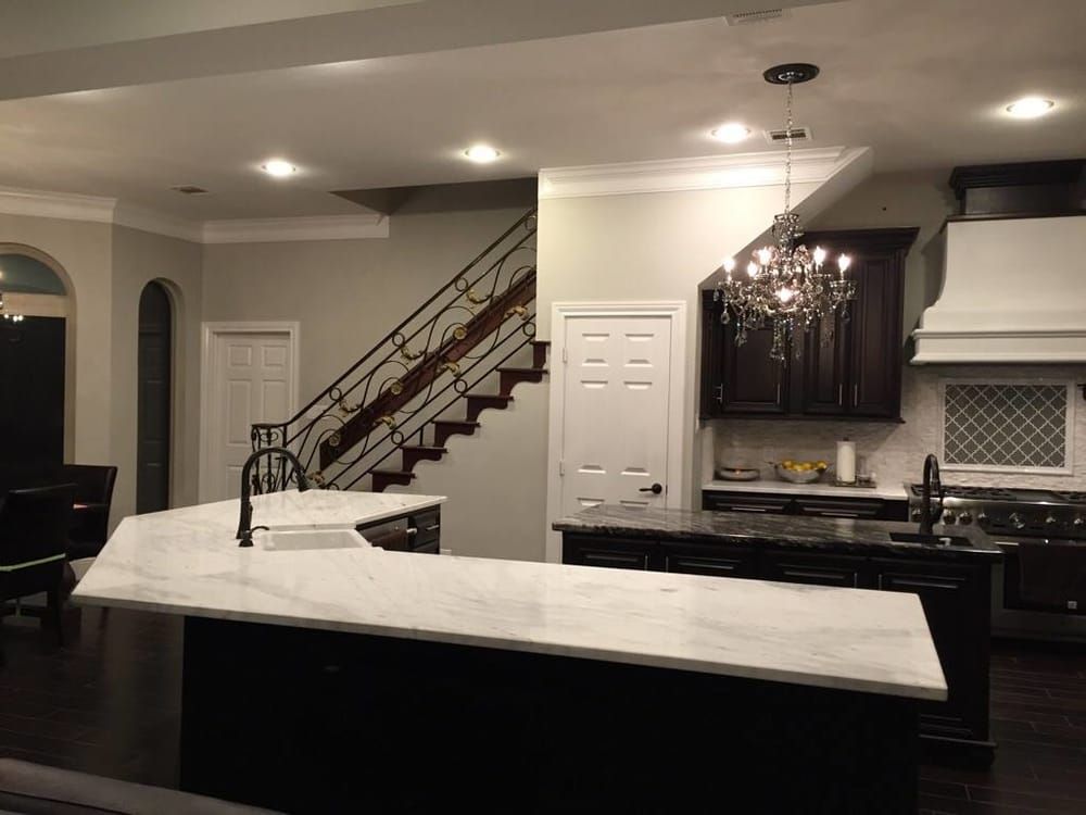 A modern kitchen featuring white marble countertops, dark cabinets, a decorative staircase, and a crystal chandelier.