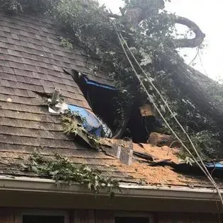 Damaged house roof with large hole, debris, and fallen tree branches after storm