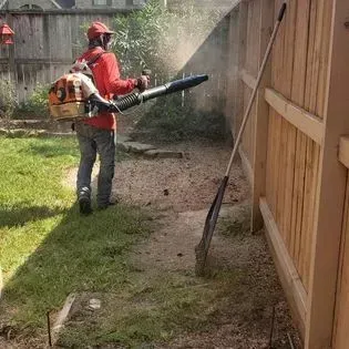 Worker using a backpack leaf blower to clear debris along a wooden fence in a yard