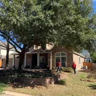 Two people in red shirts walking outside a brick house under large trees