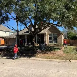 Two workers in red shirts outside a brick house under large trees, with a street in front
