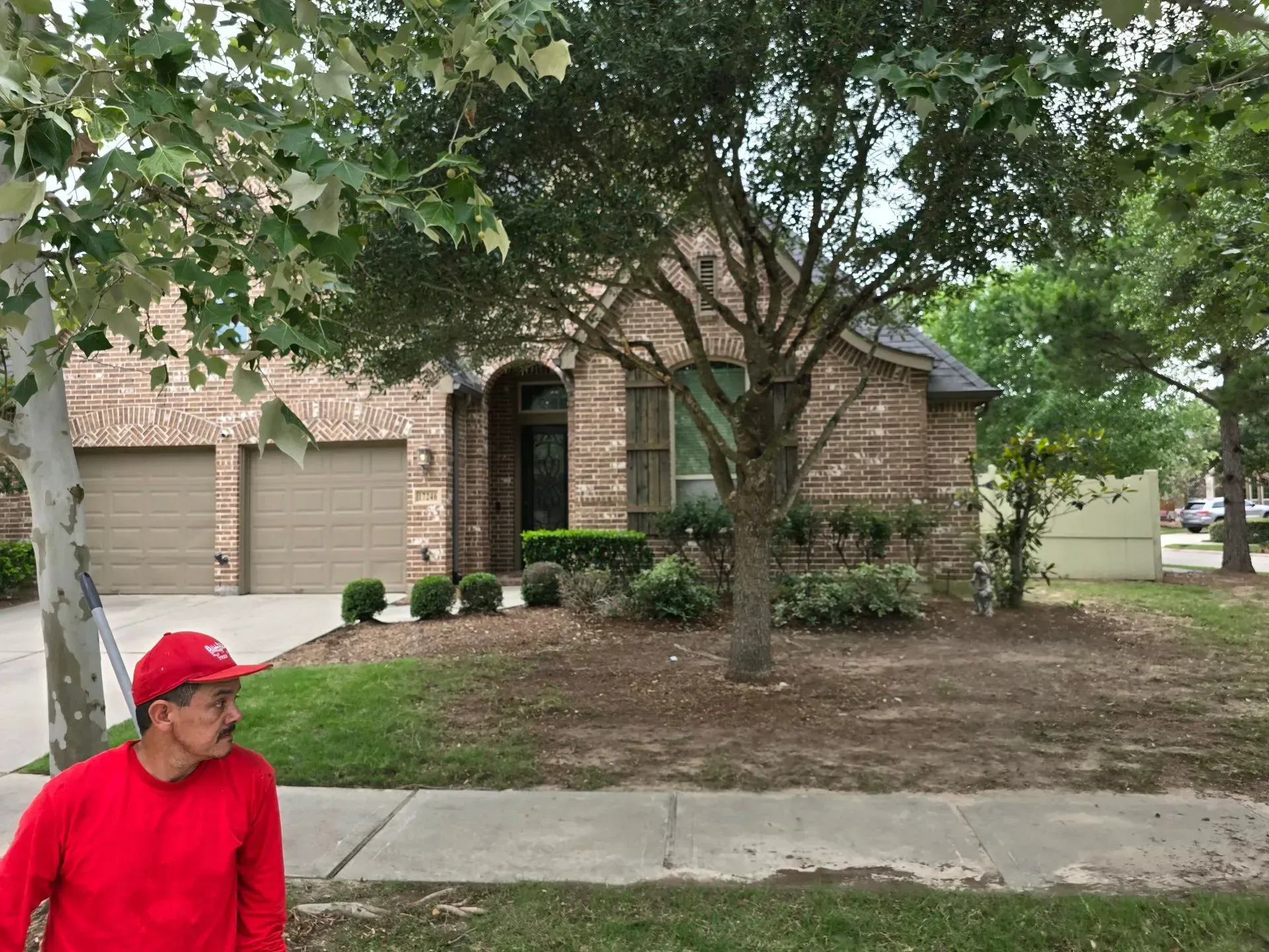 Red-shirted person standing on a sidewalk in front of a brick house with a garage and trees.