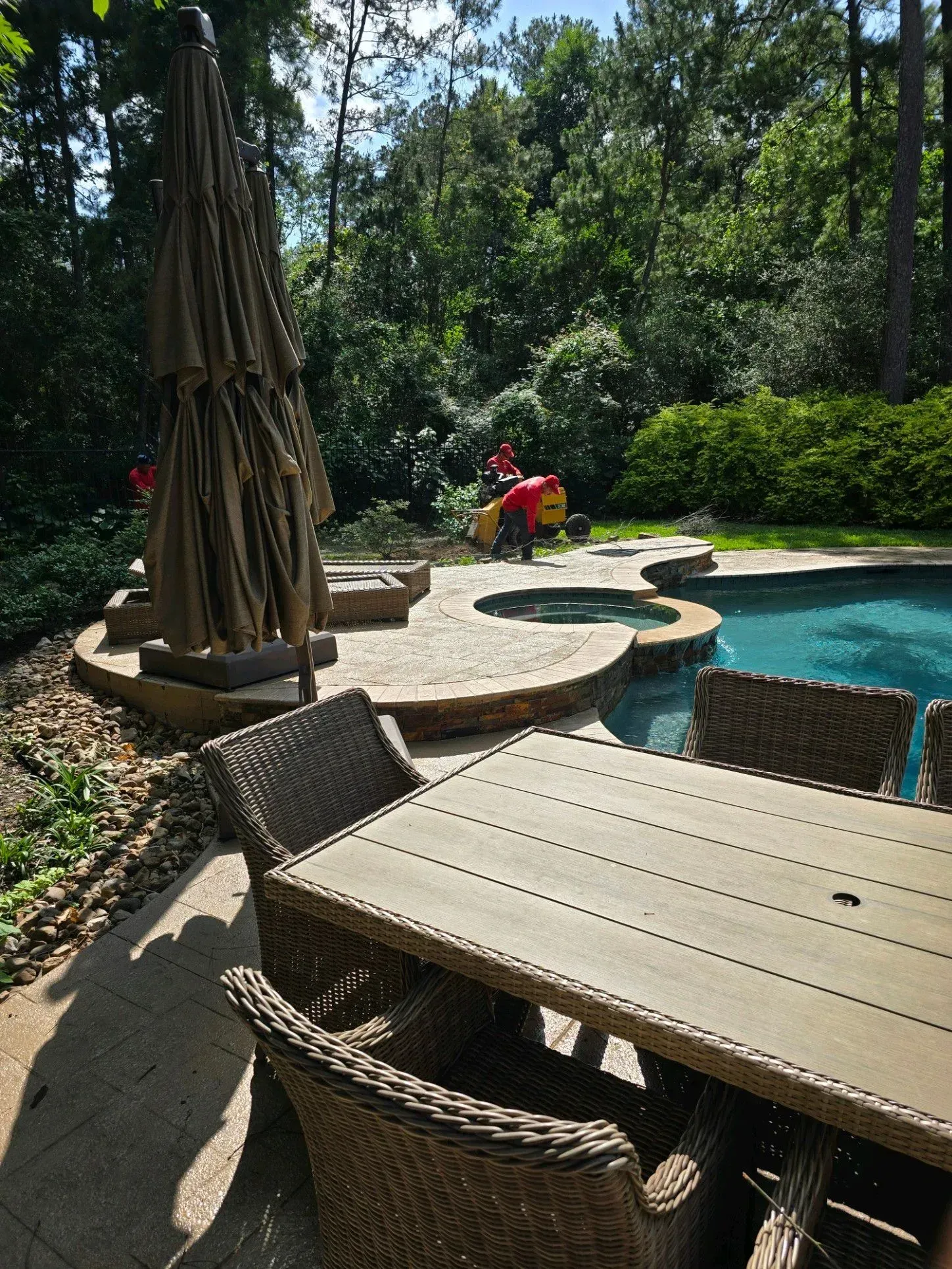 A table and chairs are sitting next to a pool. Patio table and chairs by a pool, with an umbrella and two people in the background
