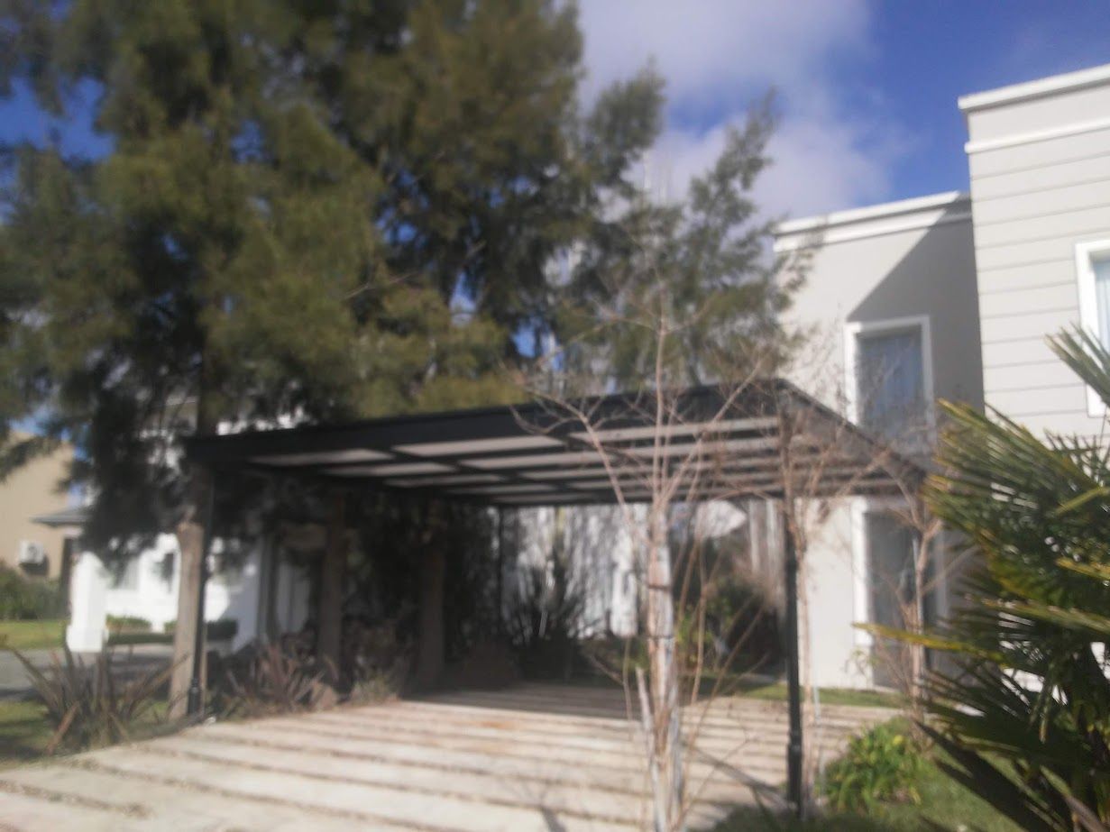 Entrada a una casa blanca con una pérgola negra sobre escalones, enmarcada por árboles y un cielo azul claro.