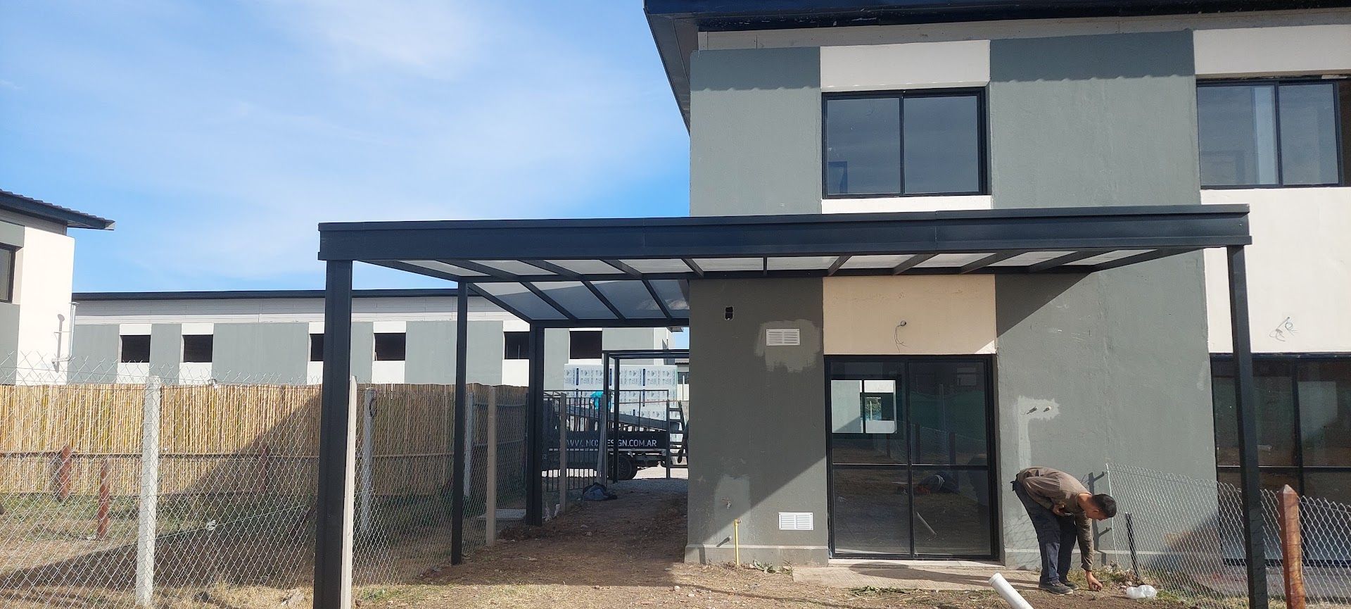 Exterior de una casa moderna con cochera, patio cercado y una persona agachada cerca de la puerta principal.